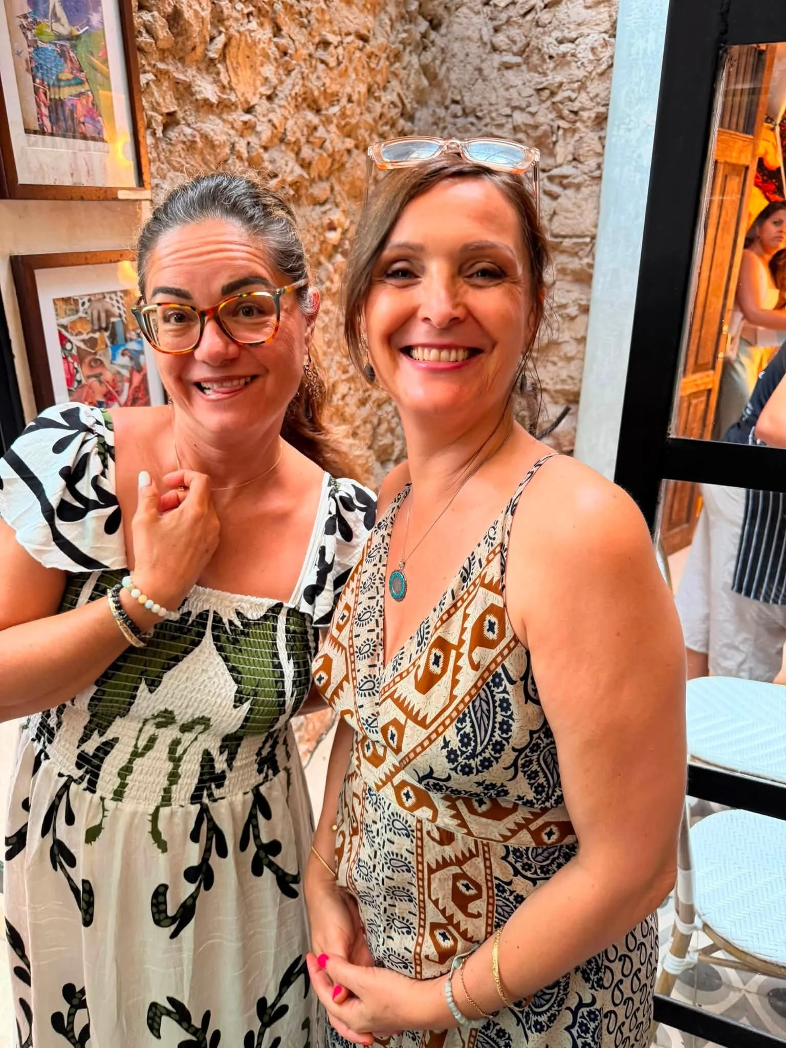 Two women smiling and posing for a photo indoors against a brick wall, with framed artwork, in a casual setting.