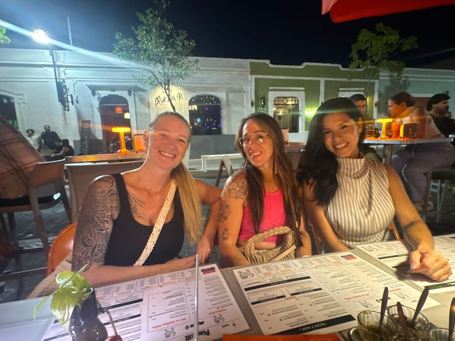 Three women smiling and sitting at a restaurant table at night, with menus in front of them and other diners in the background.
