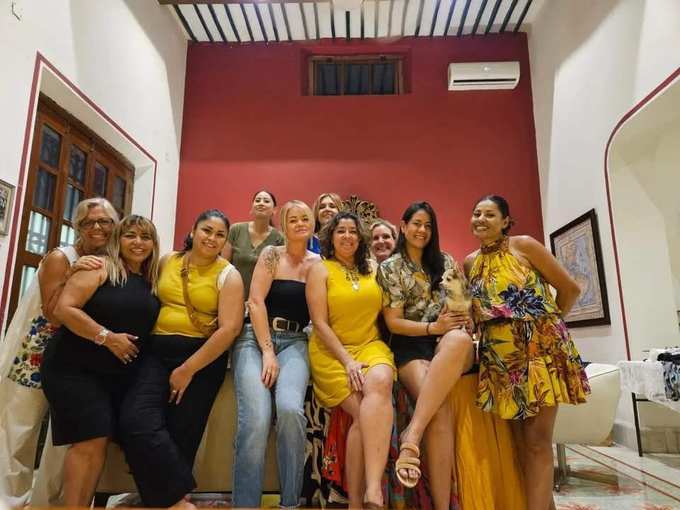 Group of ten women smiling and posing indoors, some holding a small dog, with red and white walls and framed art in the background.