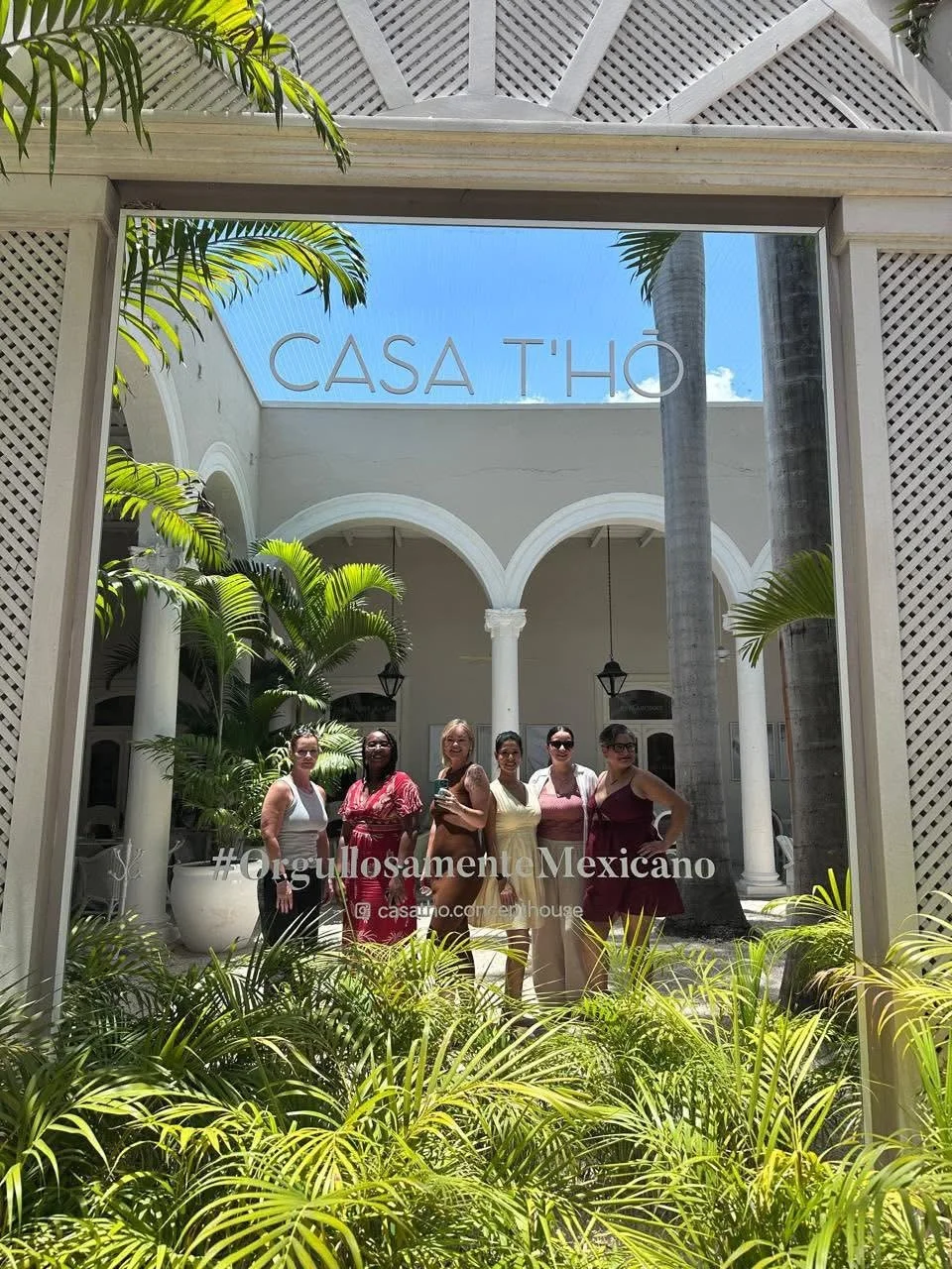 Group of women standing inside a courtyard with white columns and arches, lush green plants in the foreground, viewed through a glass. The words CASA T'HO are written on the glass.