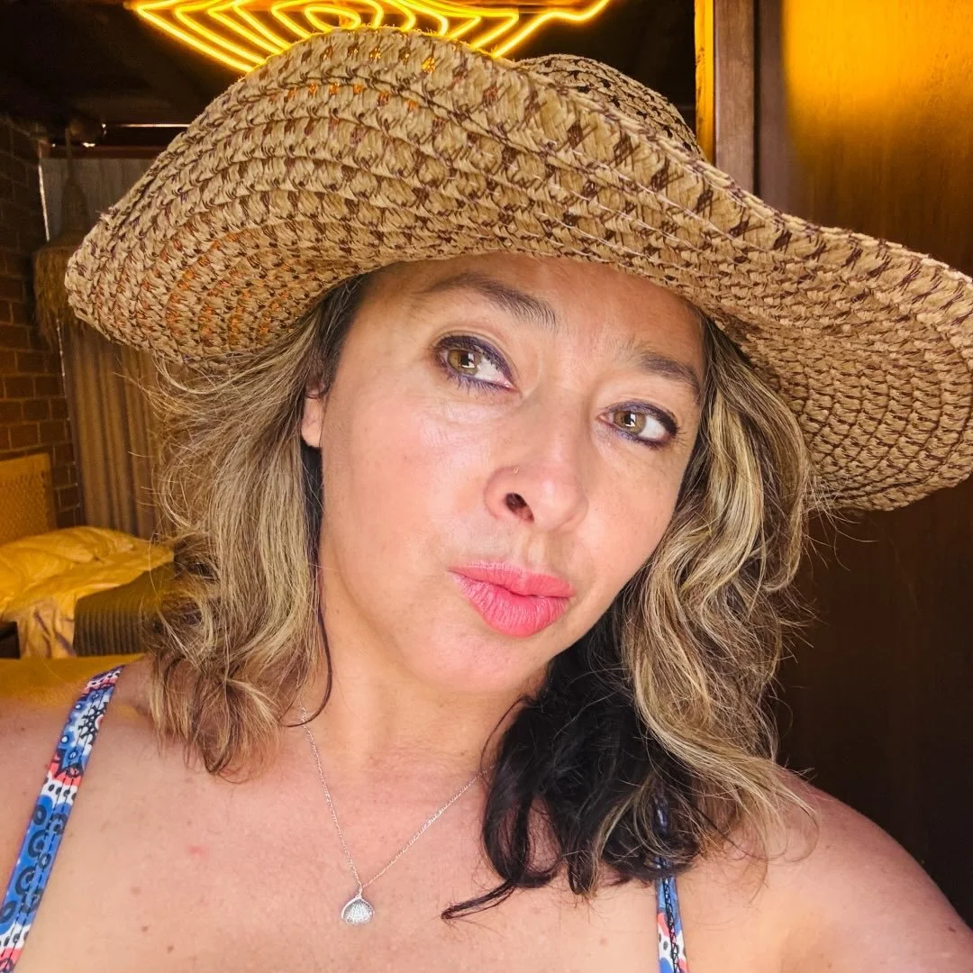 A woman with wavy light brown hair, wearing a large straw hat, a necklace with a small pendant, and a blue patterned tank top, sitting indoors with a bed and brick wall in the background.
