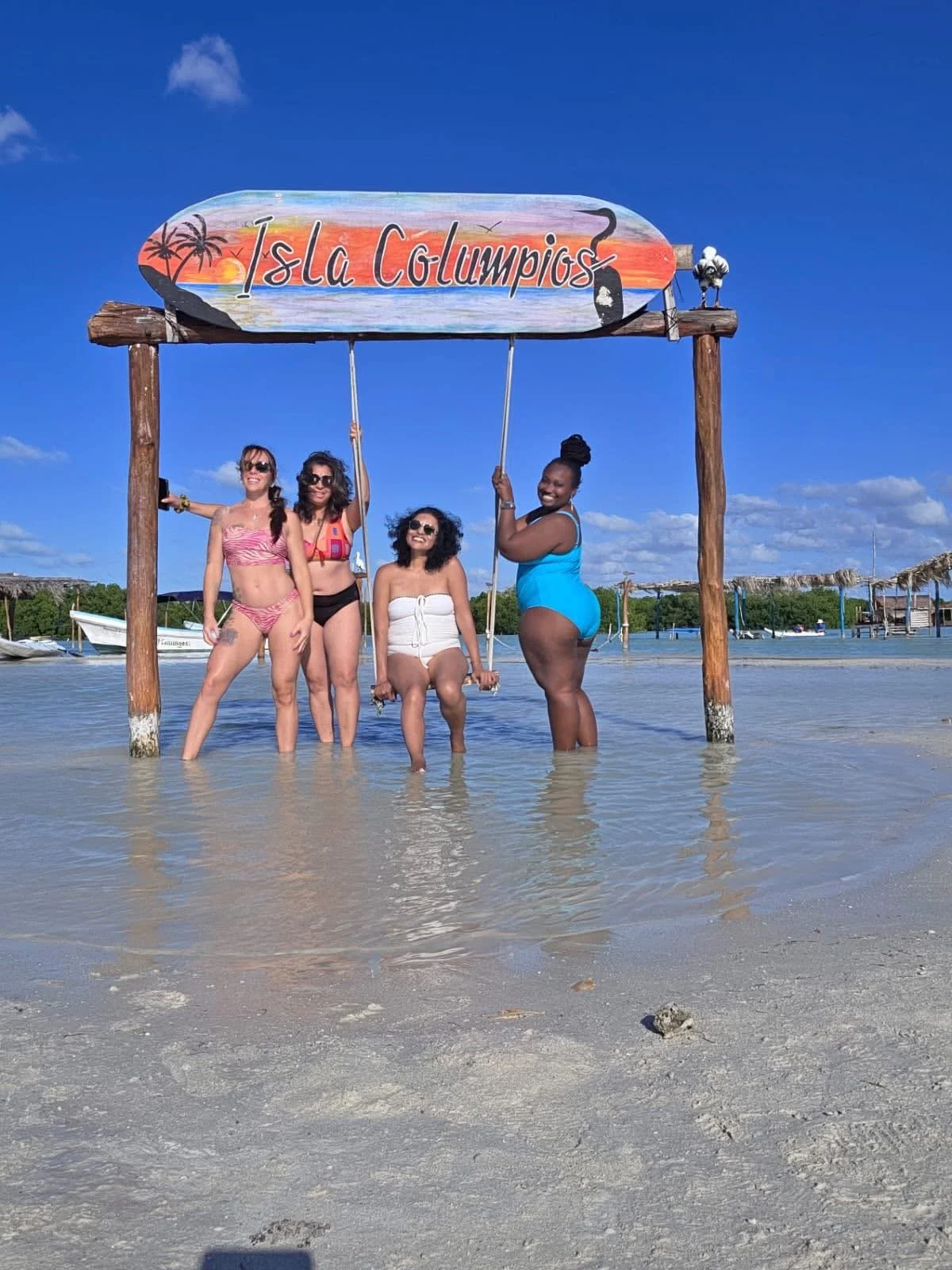 Four women in swimsuits enjoy the shallow water at Isla Columpios, a tropical island with clear blue skies, boats in the background, and a colorful sign overhead.