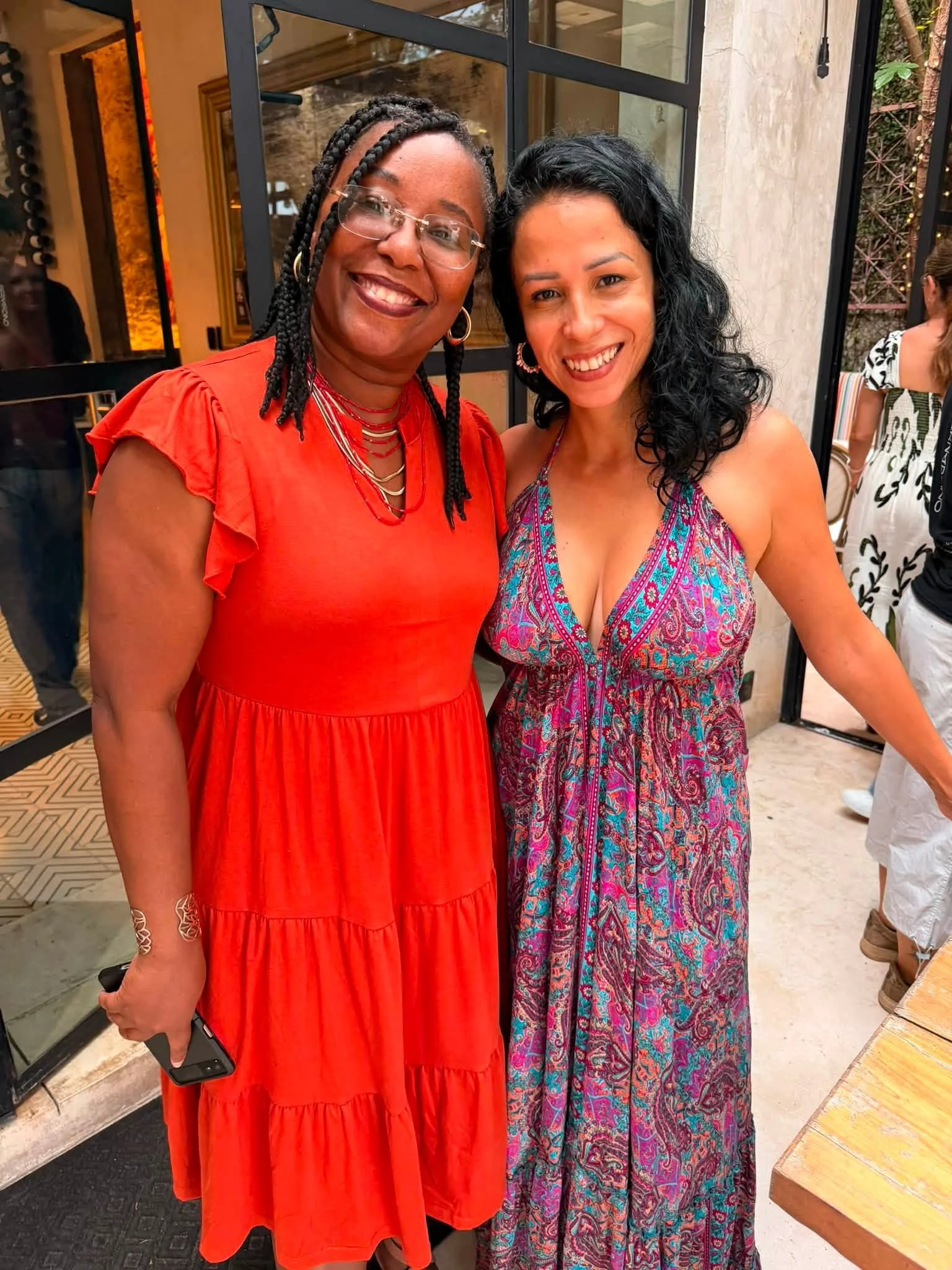 Two women smiling, standing close together in a well-lit indoor setting. The woman on the left is African American, wearing glasses, a red dress with ruffled sleeves and layered necklaces. The woman on the right is of mixed ethnicity, with curly blac