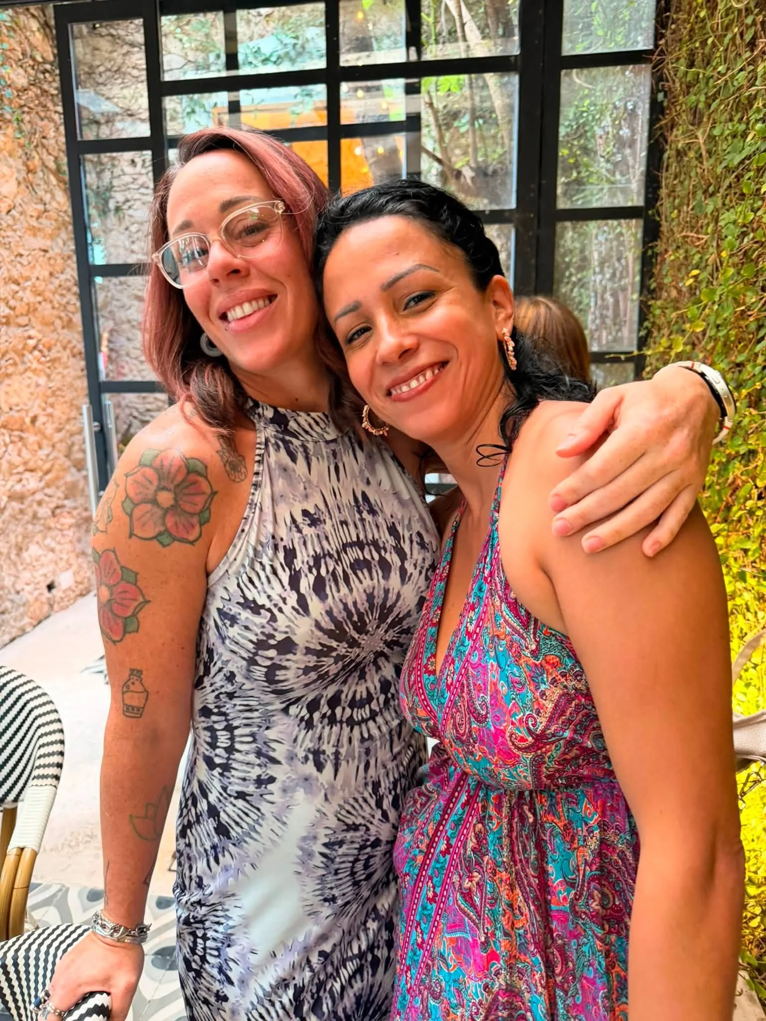 Two women smiling and hugging each other in an outdoor setting with large windows and greenery in the background. One woman has tattoos on her arm and wears glasses, the other has earrings and wears a colorful patterned dress.