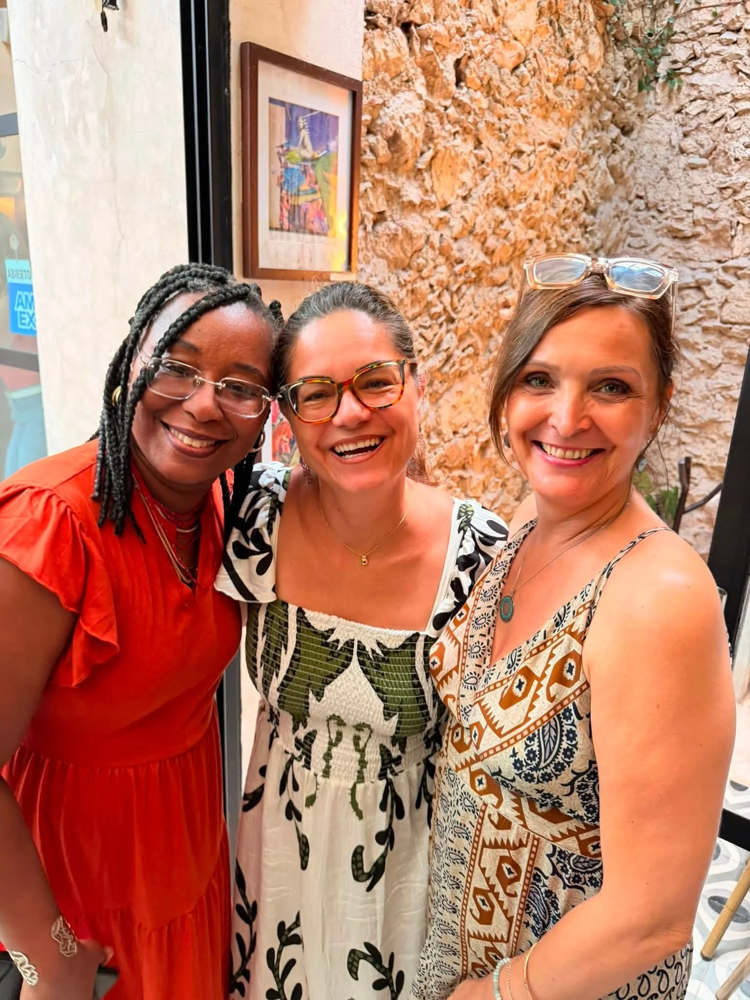 Three women smiling and posing for a photo in a lively, colorful setting with a rustic stone wall and artwork in the background. One woman has glasses and braided hair, another has dark glasses and a patterned dress, and the third has sunglasses on h