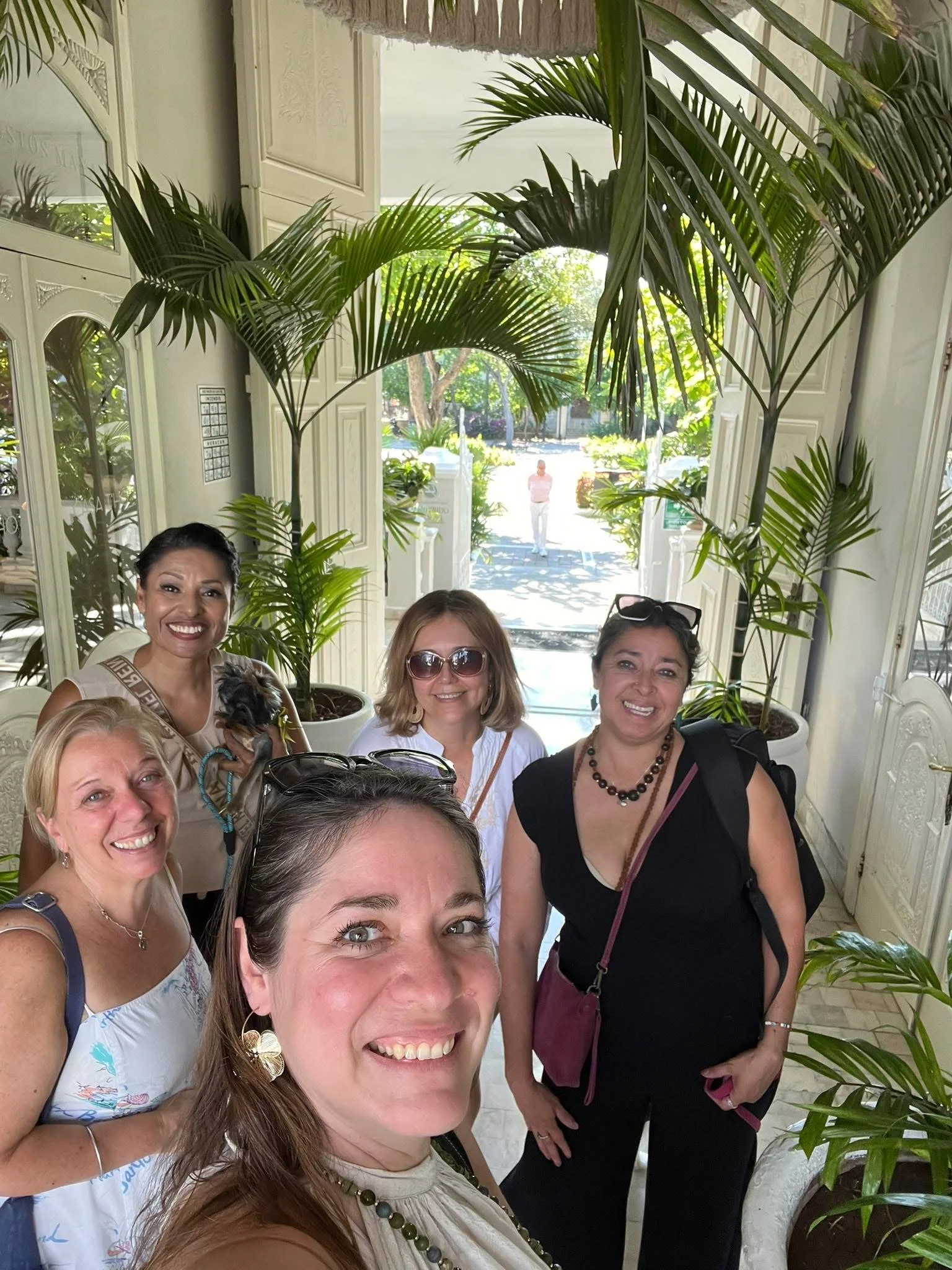 Group of smiling women taking a selfie inside a building with large potted plants and an open door showing a street with trees and a person in the background.