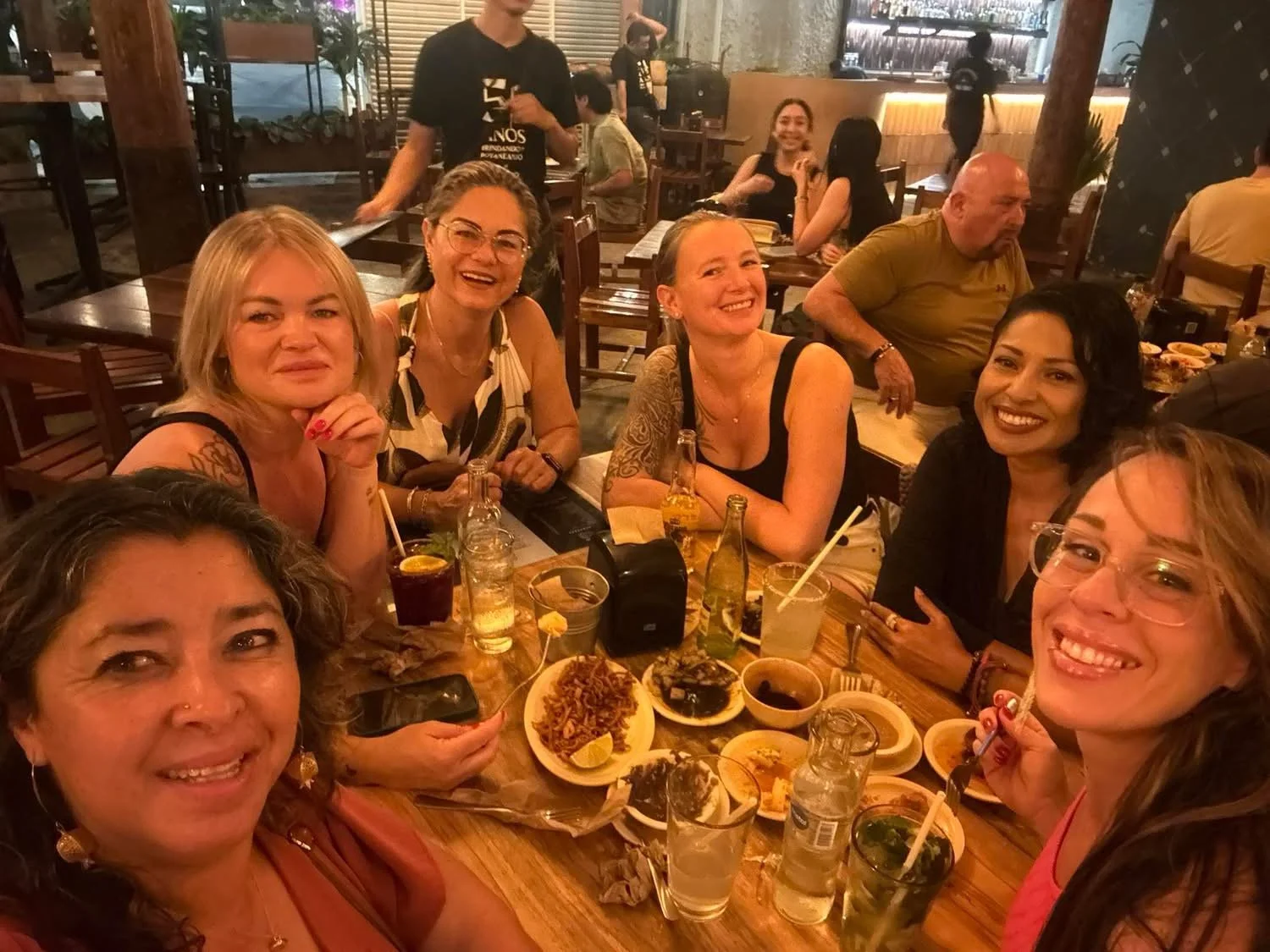 A group of women smiling and enjoying drinks and food at a restaurant table.