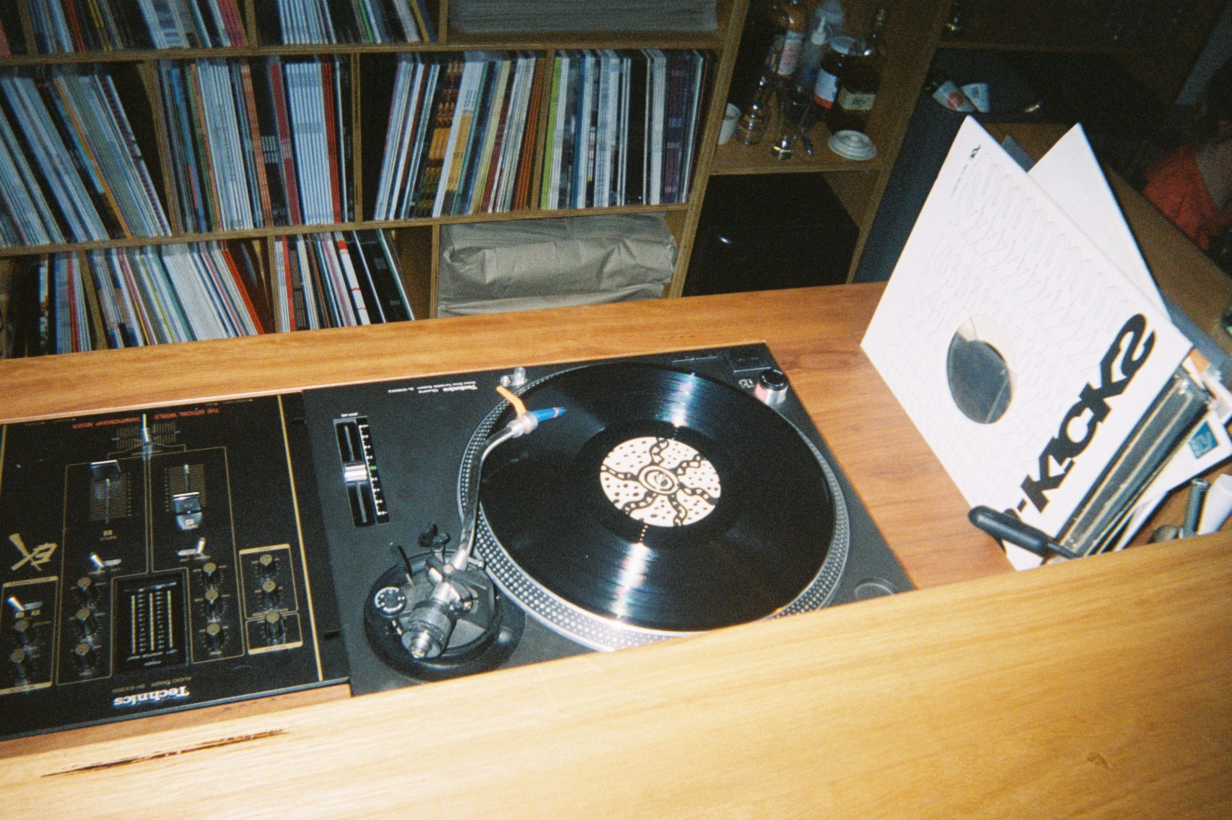 A turntable playing a vinyl record next to a white box with black and orange text, with a wooden desk and a bookshelf filled with books and magazines in the background.