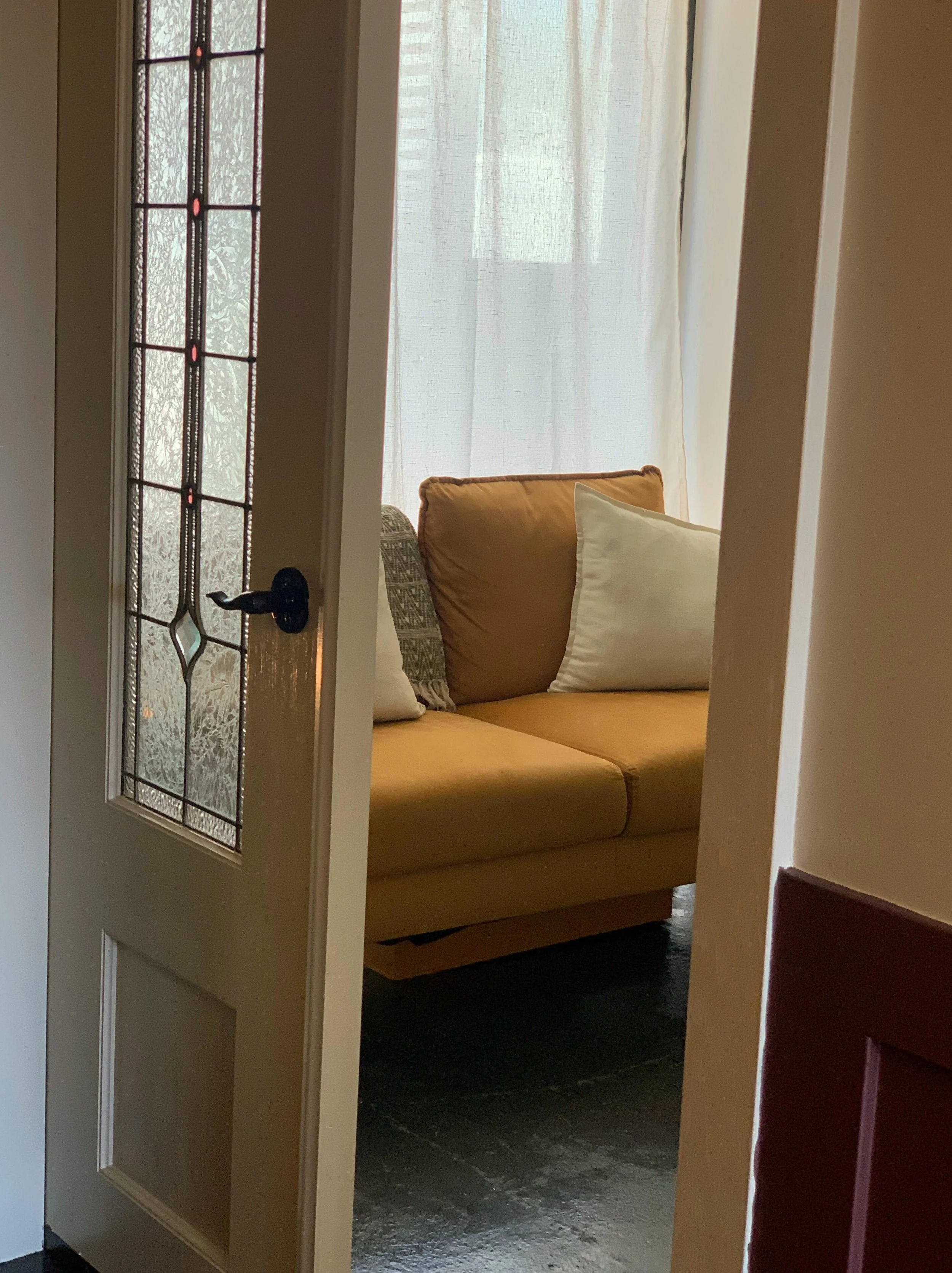 View through a partially open door into a cozy living room with a yellow sofa, decorative pillows, sheer white curtains, and a window letting in natural light.