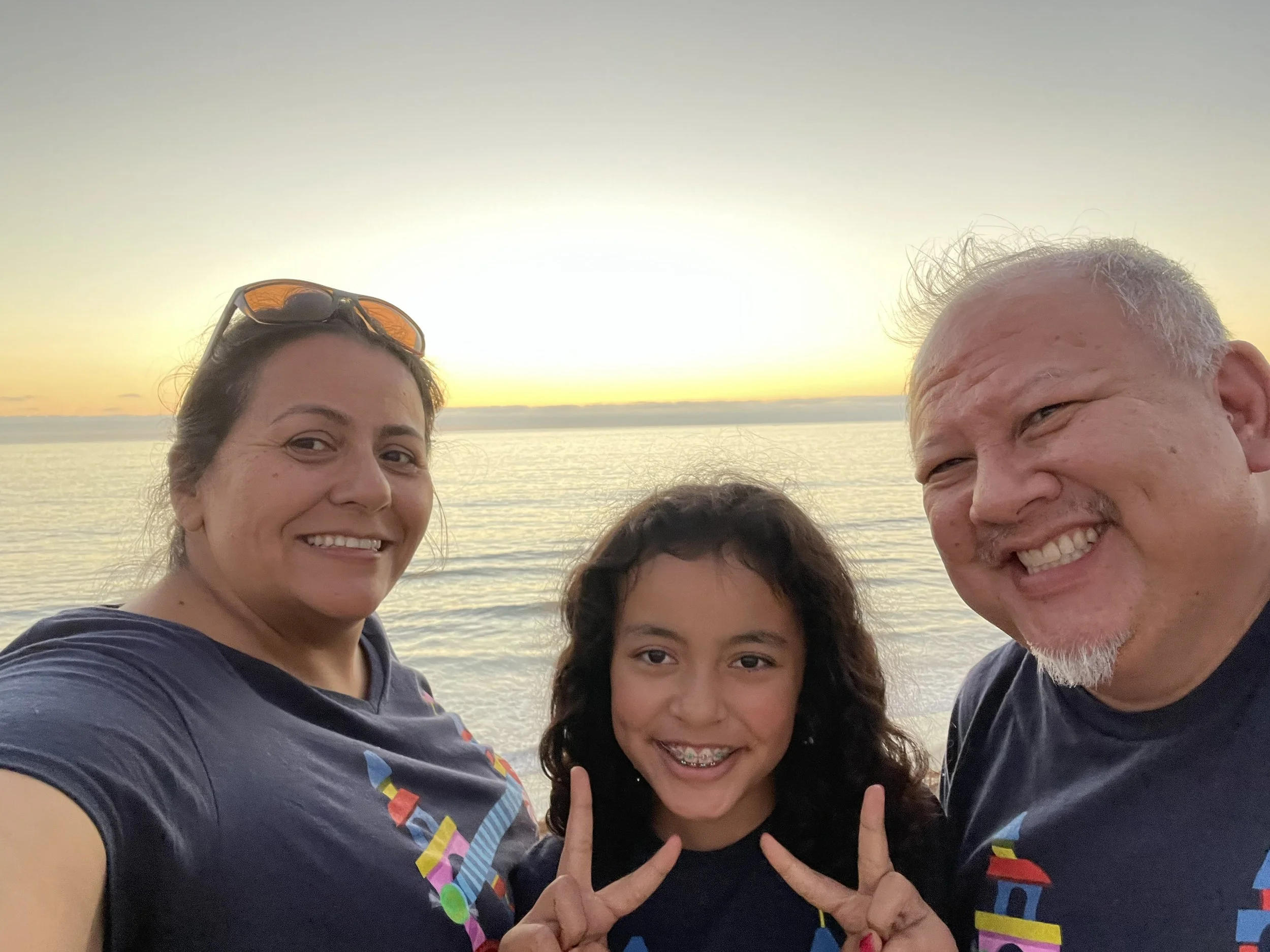 Group of four people smiling by the water during sunset, with one person making a peace sign.