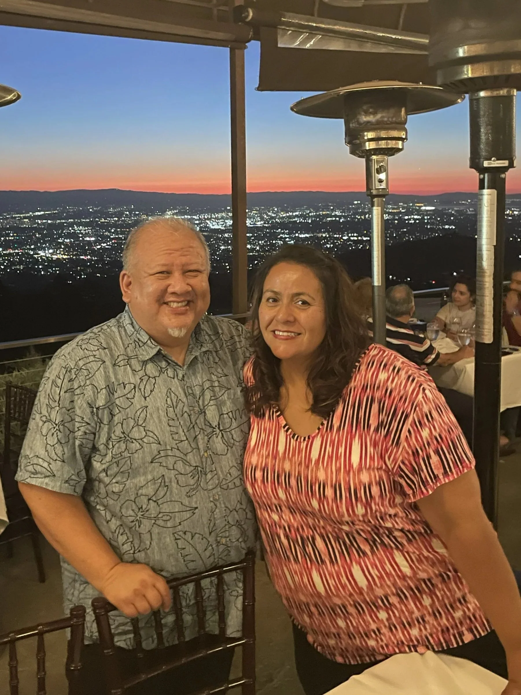 Smiling man and woman standing together at an outdoor restaurant with city lights and a sunset in the background.