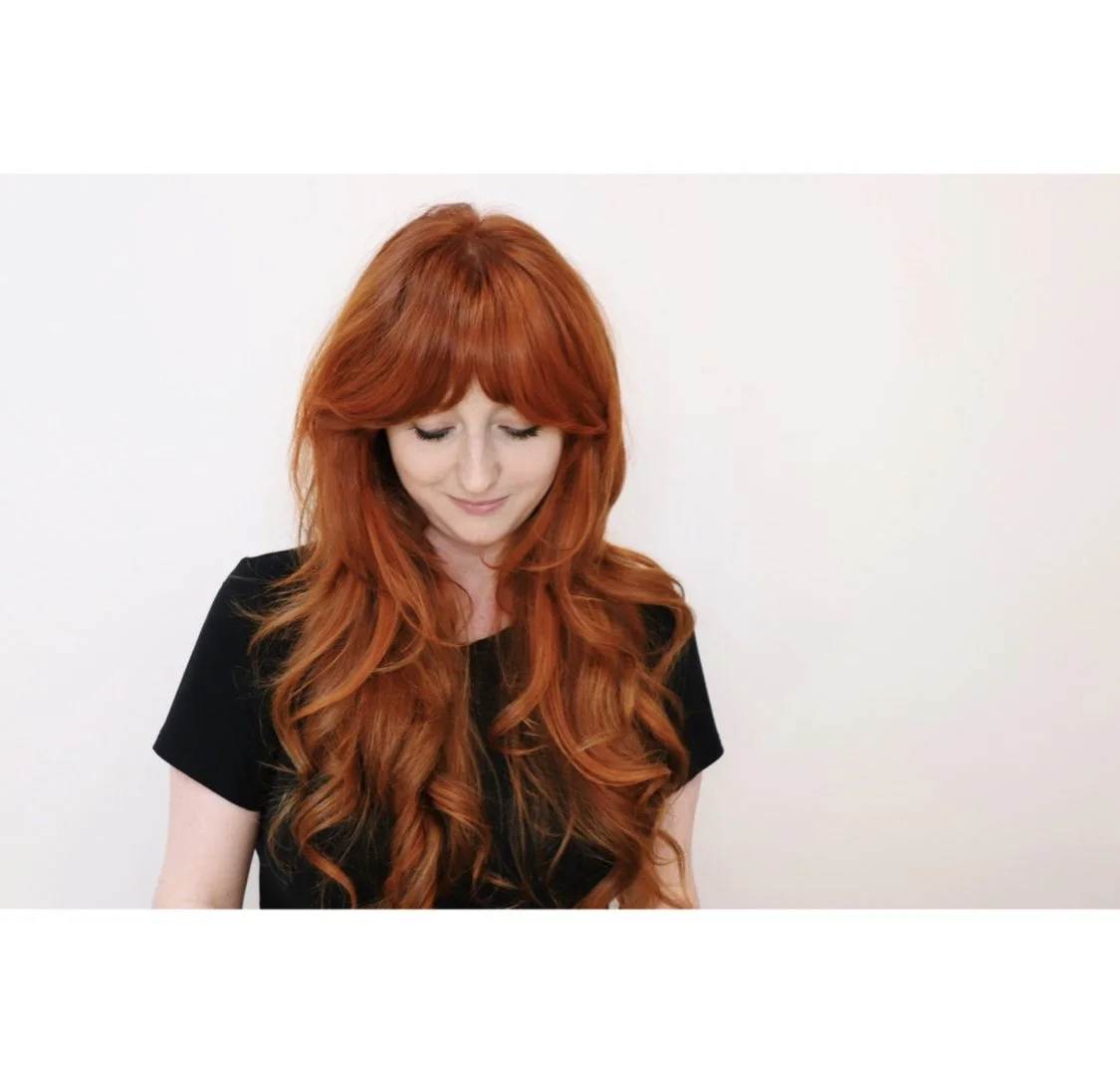 Woman with long red hair and black top looking down against a white background.