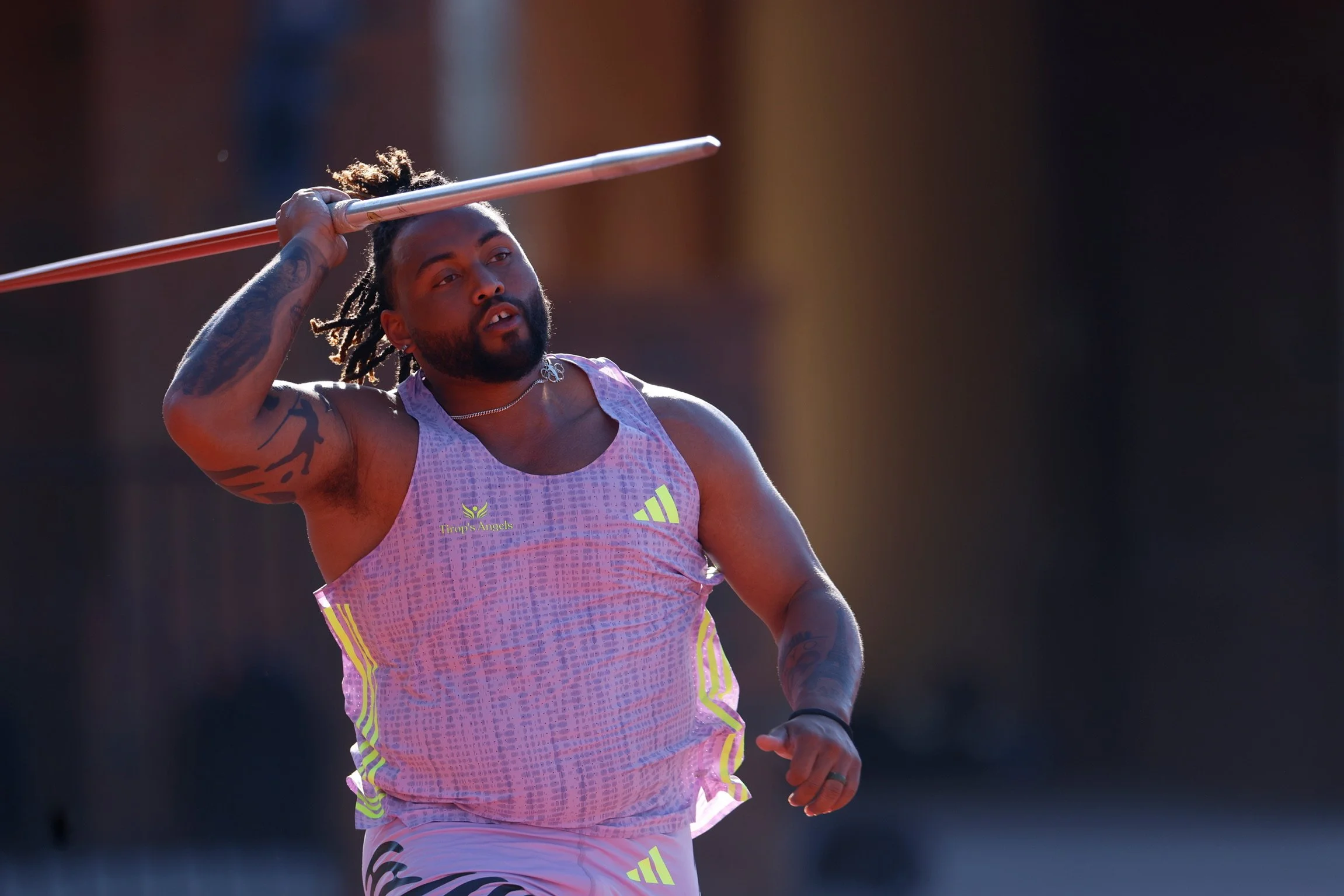 Athlete with dreadlocks lifting a javelin during a track and field event, wearing a pink and yellow uniform with 'Firep's Angels' logo.
