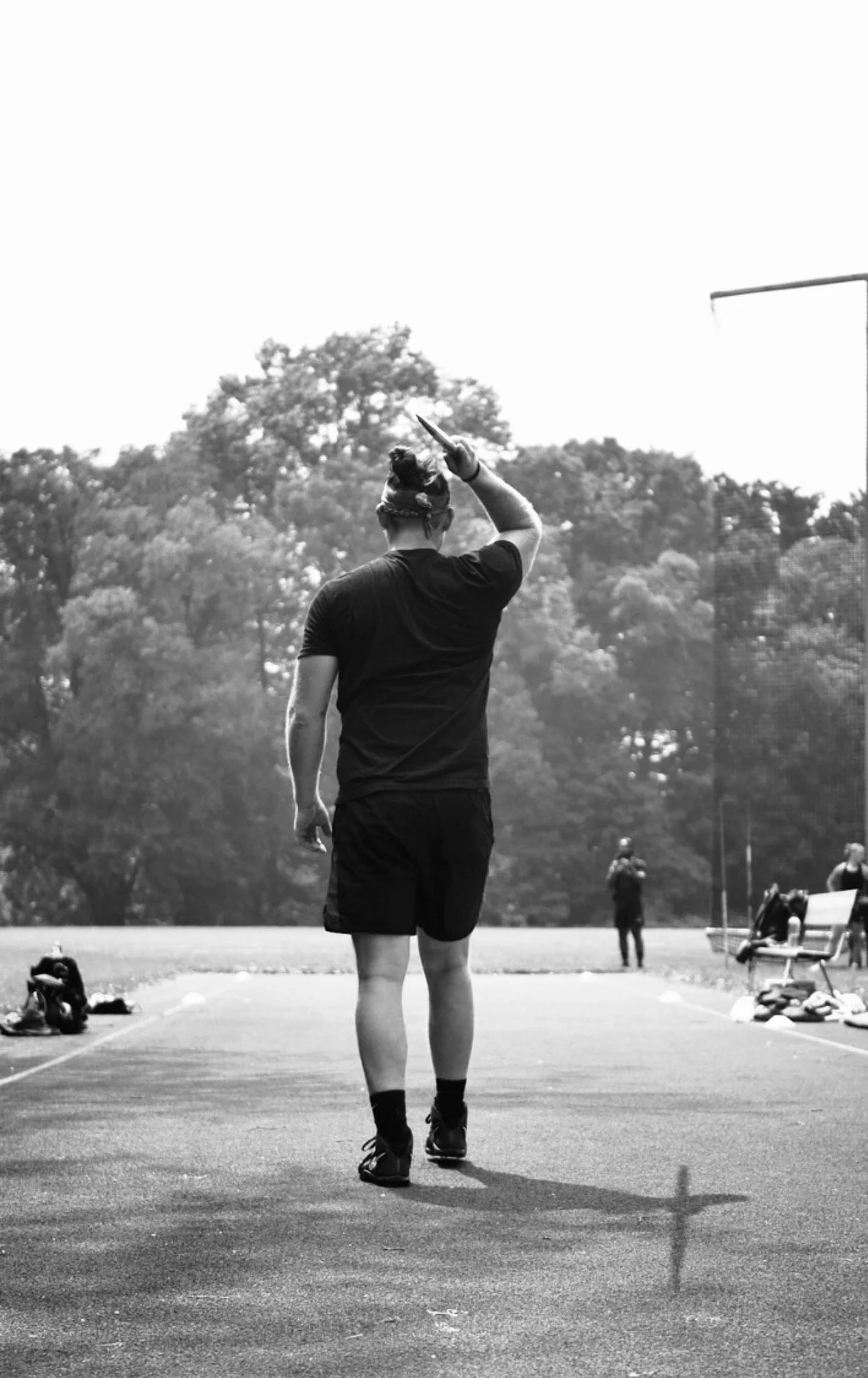 A person standing on a track field, seen from behind, holding a baton above their head. Other people are visible in the background, with some sitting on benches and others standing, with trees beyond the track.