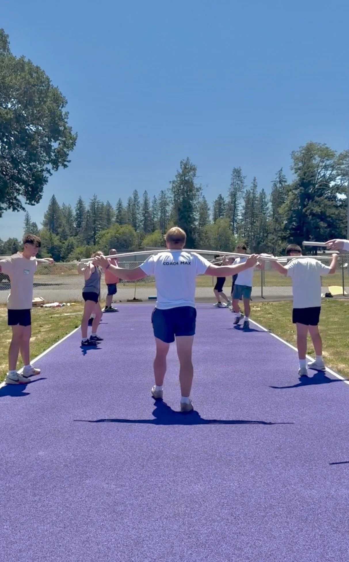 Group of people doing warm-up exercises outside on a blue sports court on a sunny day, with trees and a clear sky in the background.