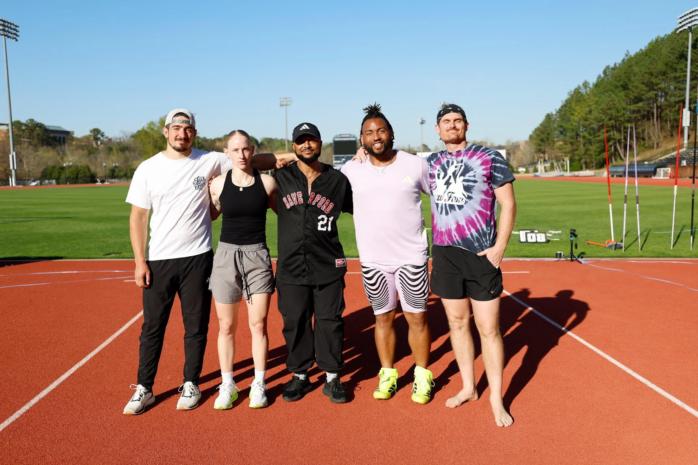 Group of five runners standing on a track field, with three men and two women, outdoors on a sunny day. Some are wearing athletic gear, one woman is barefoot, and they are smiling at the camera. There are stadium lights and trees in the background.