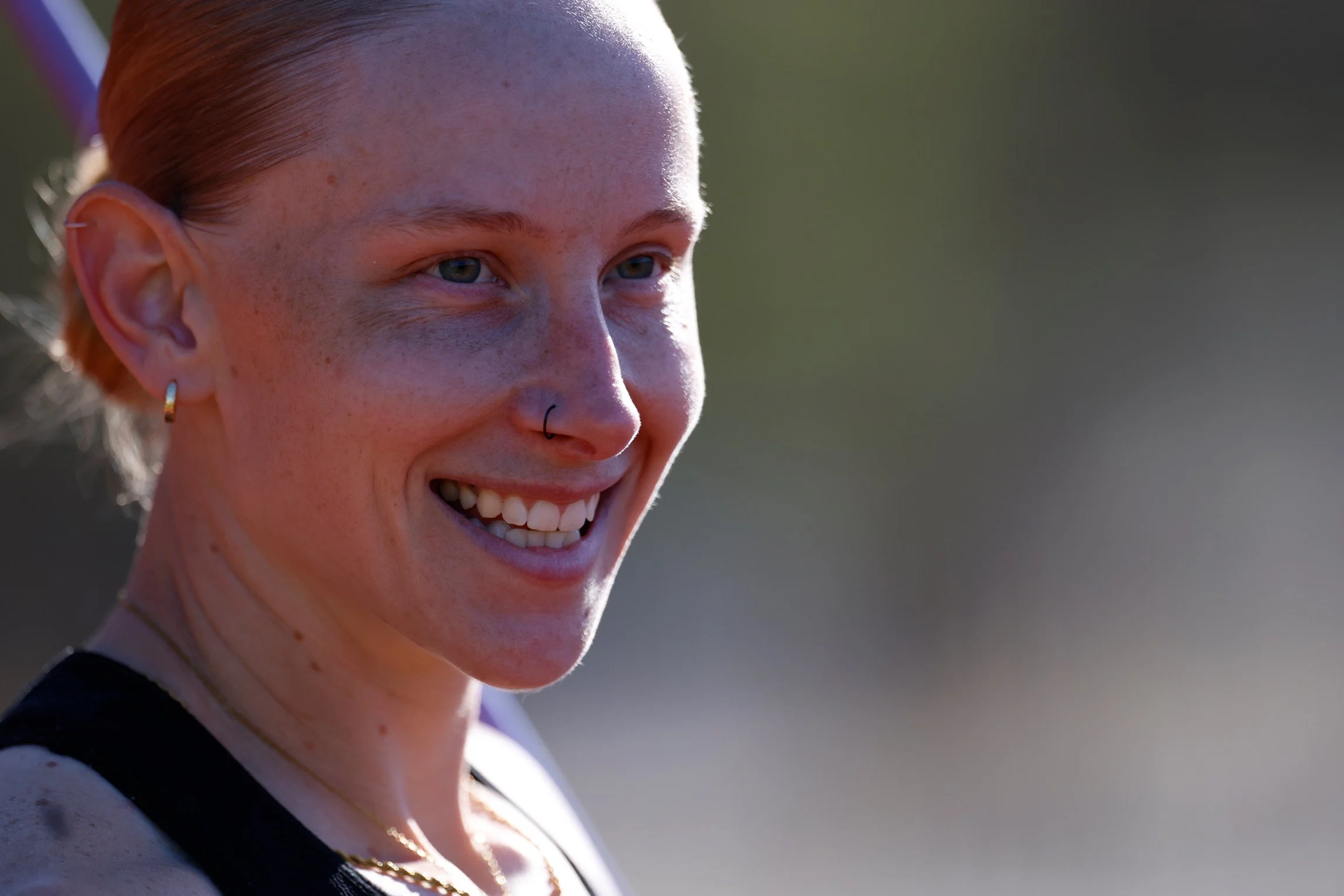 Close-up of a smiling woman with freckles and red hair tied back, wearing a nose piercing and a gold earring, outdoors in natural light.