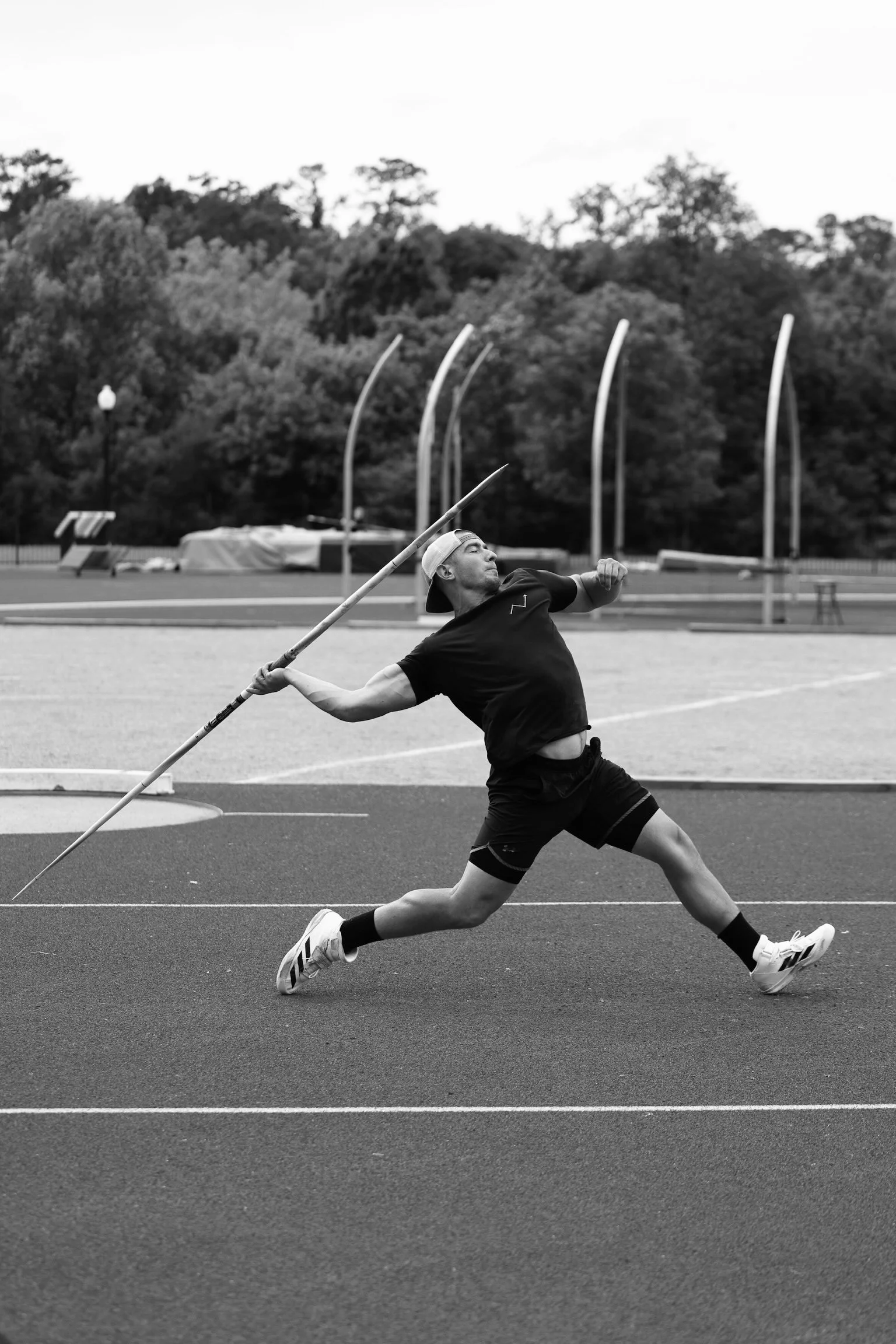 A male athlete in black sportswear and a backwards cap throws a javelin on a track field with trees and stadium equipment in the background.