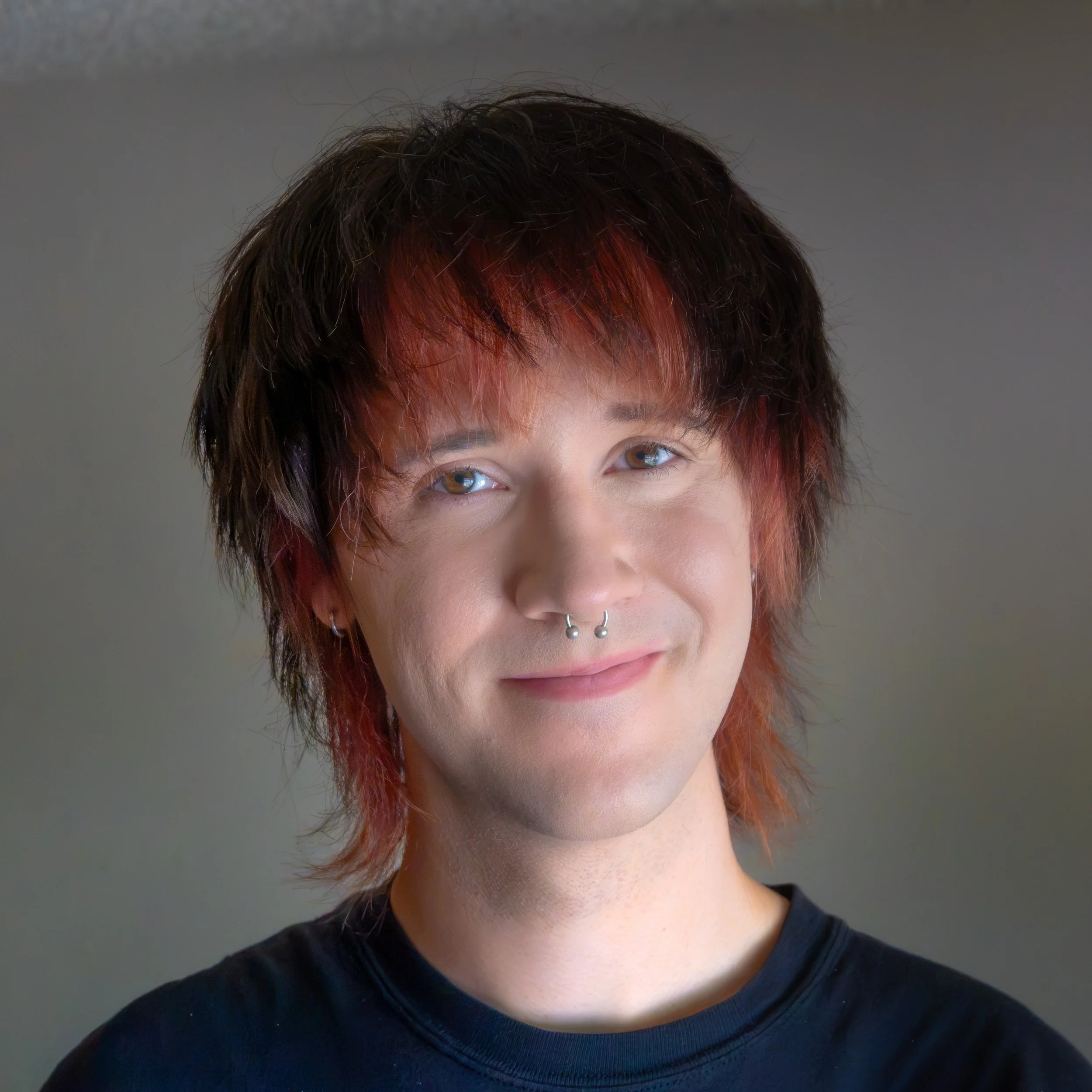 Close-up of a person with short, multi-colored hair that is black with red highlights at the front, wearing a septum piercing, and a black shirt.