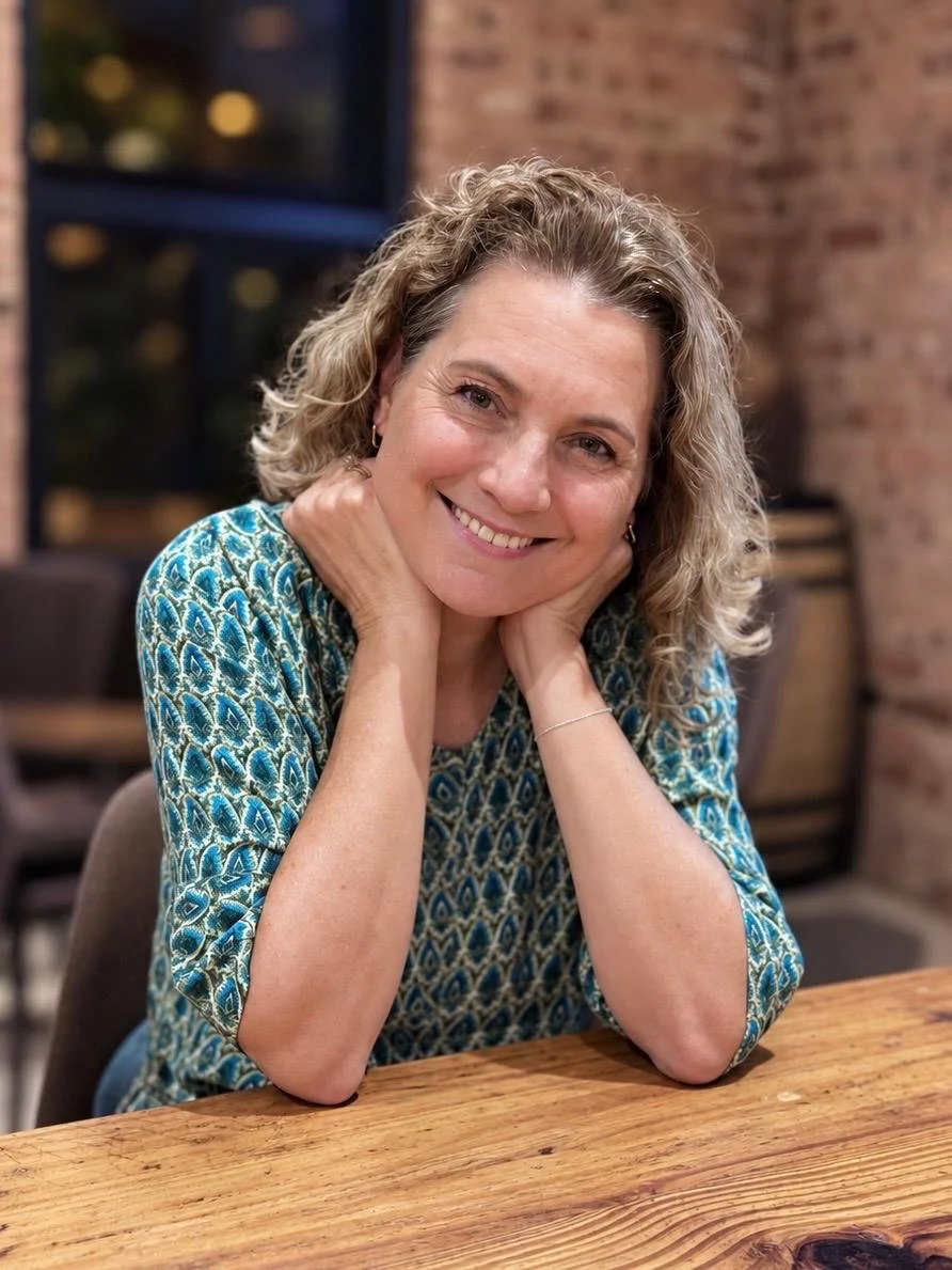 A woman with curly blonde hair smiling and resting her chin on her hands, sitting at a wooden table in a cozy indoor setting with brick walls. warm and inviting counsellor.