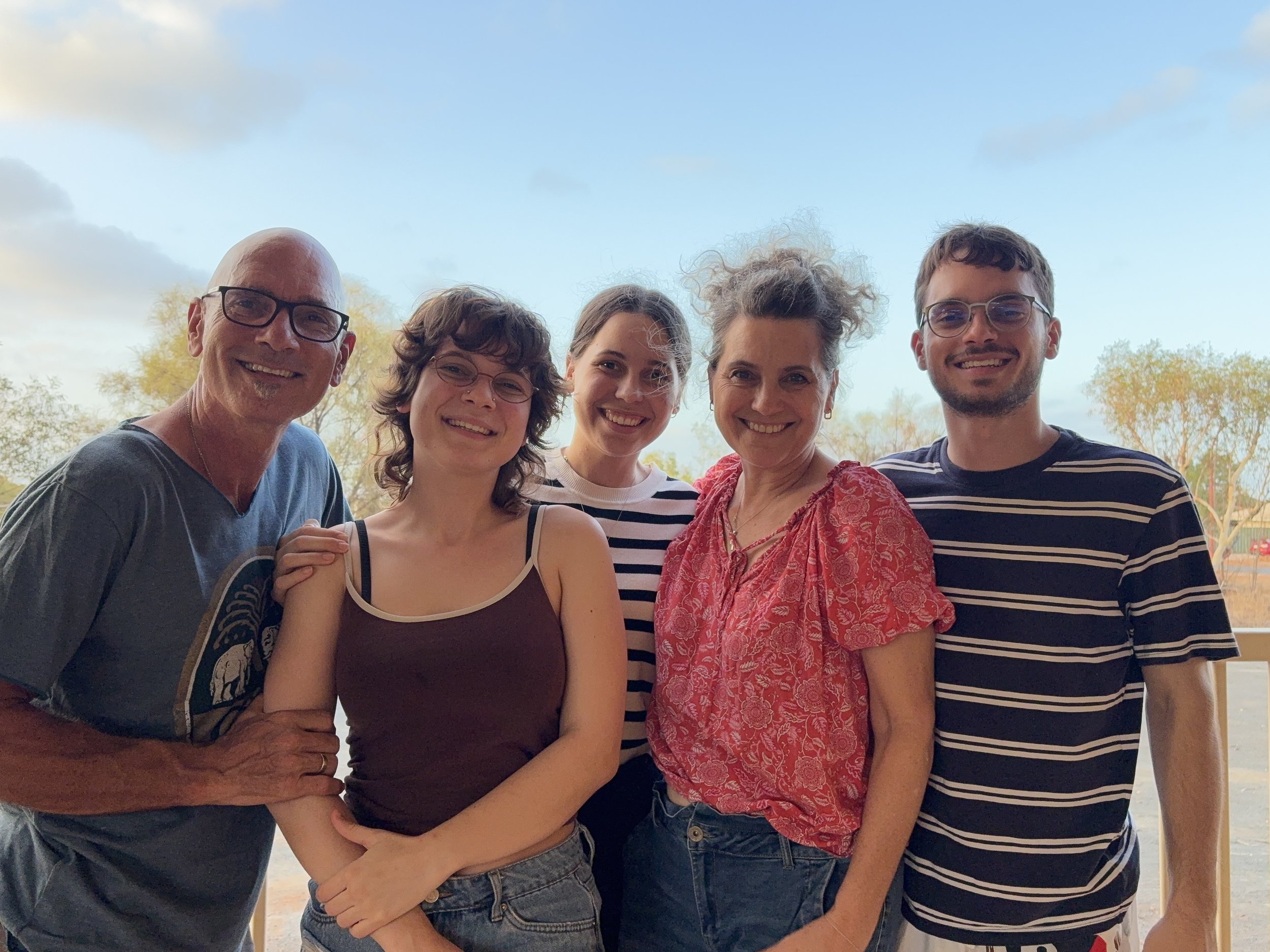 Happy family of five - parents and grown children standing outdoors in Karratha, smiling, with trees and a blue sky in the background.