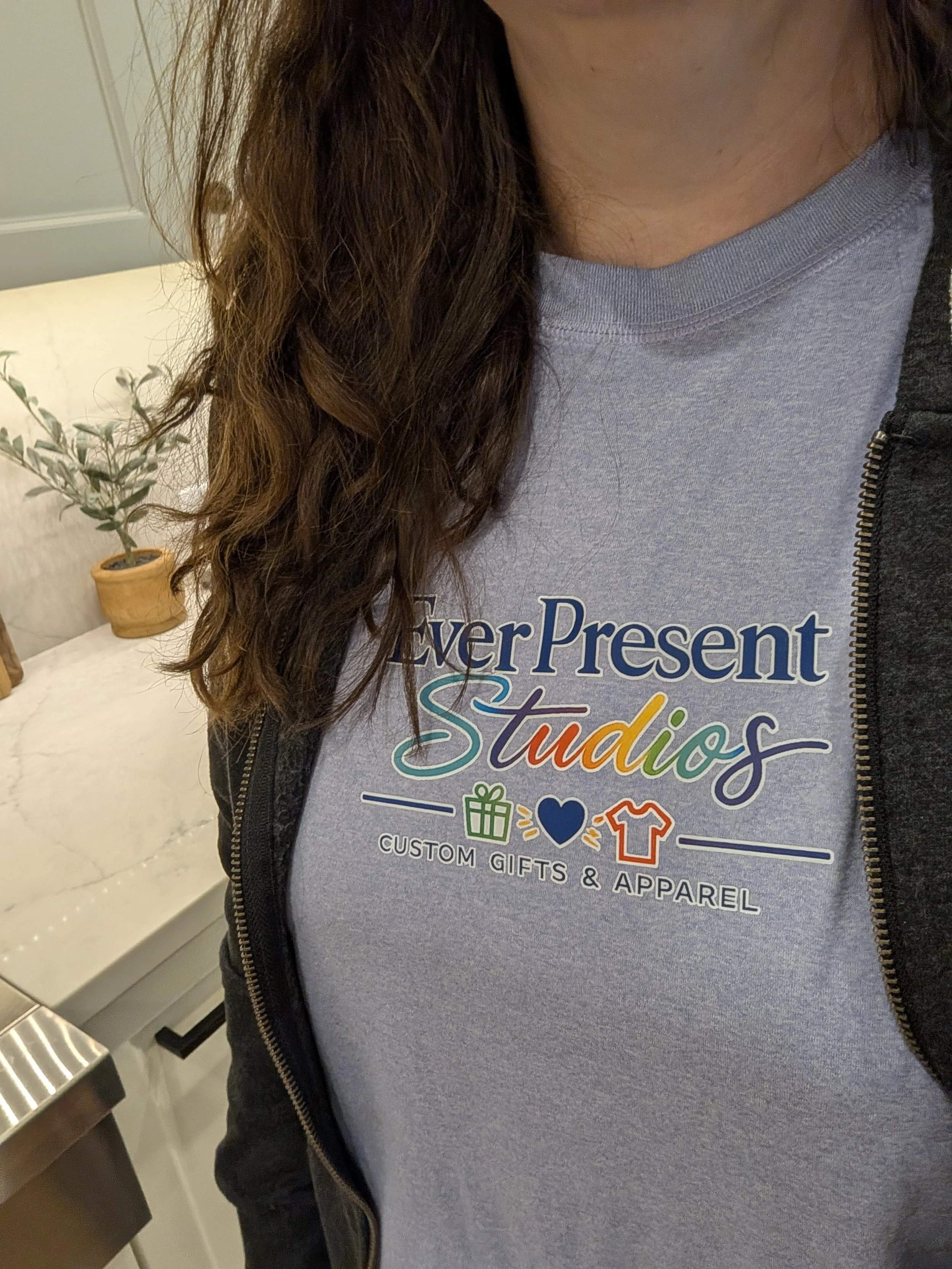 Person wearing an Ever Present Brand blue t-shirt with colorful text and graphics, partially covered by a grey zip-up hoodie, with a potted plant in the background.