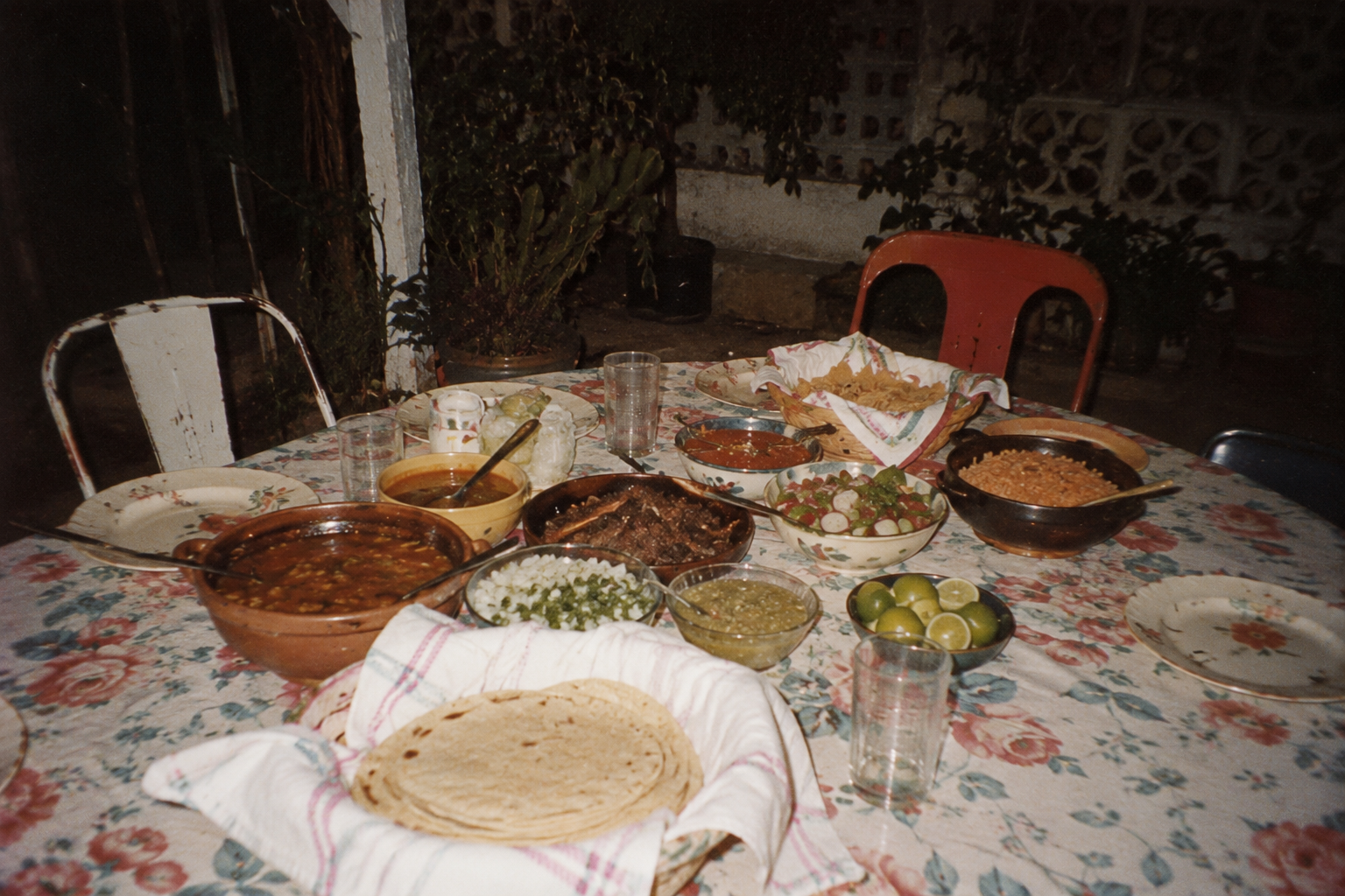 A dinner table set with various Mexican dishes, including tortillas, beans, soups, grilled meat, rice, lime wedges, and bowls of salsa, on a floral tablecloth with glass of water and empty plates.