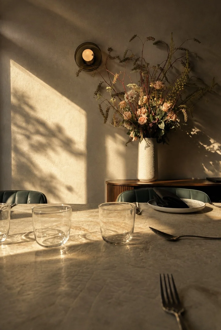 Sunlit dining table set with glasses, a spoon, and black napkins; in the background, a tall vase with pink flowers and greenery on a wooden sideboard, with a wall-mounted light and shadows from sunlight.