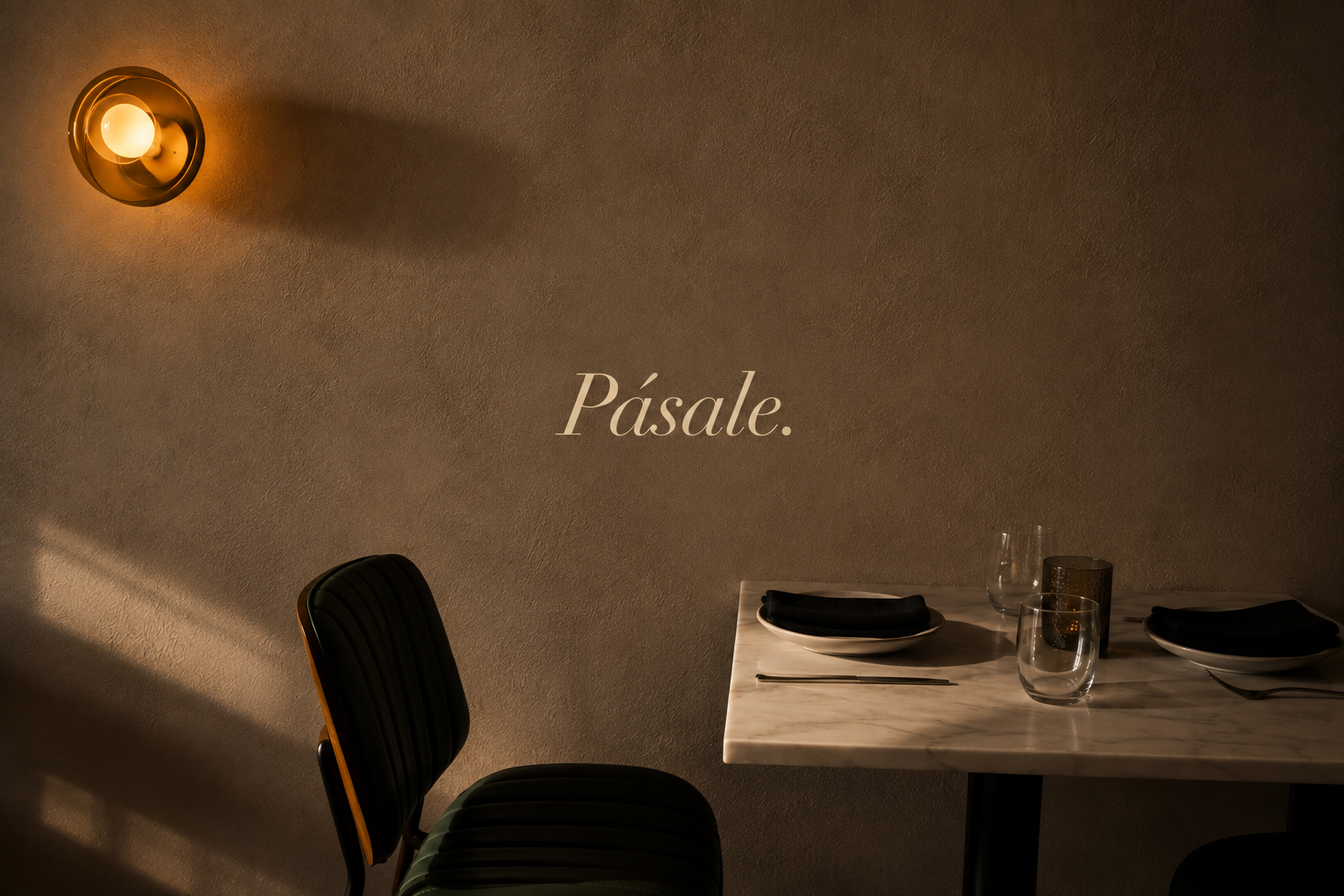 A dimly lit dining room with a modern wall-mounted light fixture, a marble table set with black napkins, white plates, wine glasses, and a candle holder, with the word 'Pásale.' written on the wall.