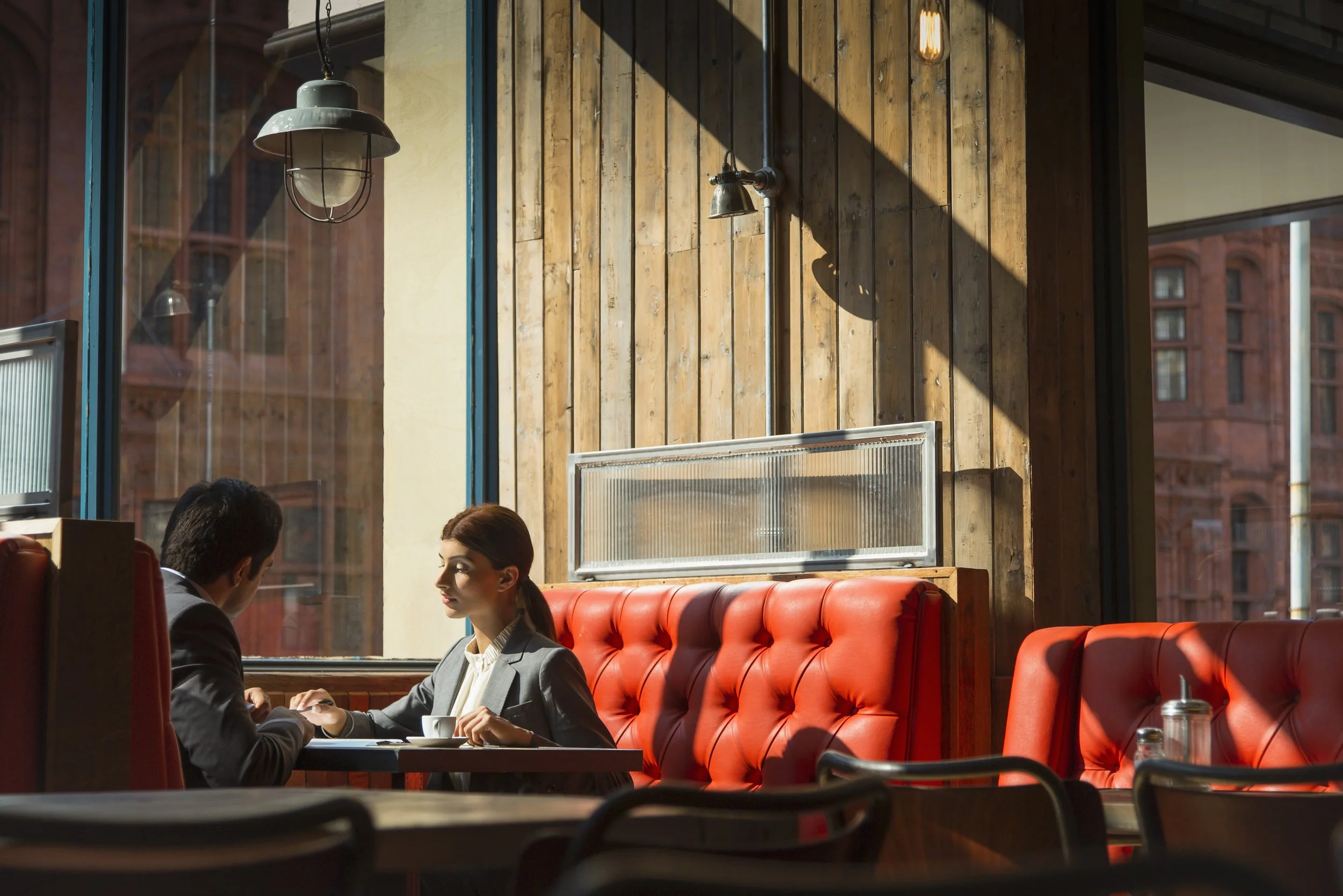 Two people sitting in a booth at a restaurant, with a large window and wooden wall behind them, one woman and one man engaging in conversation.