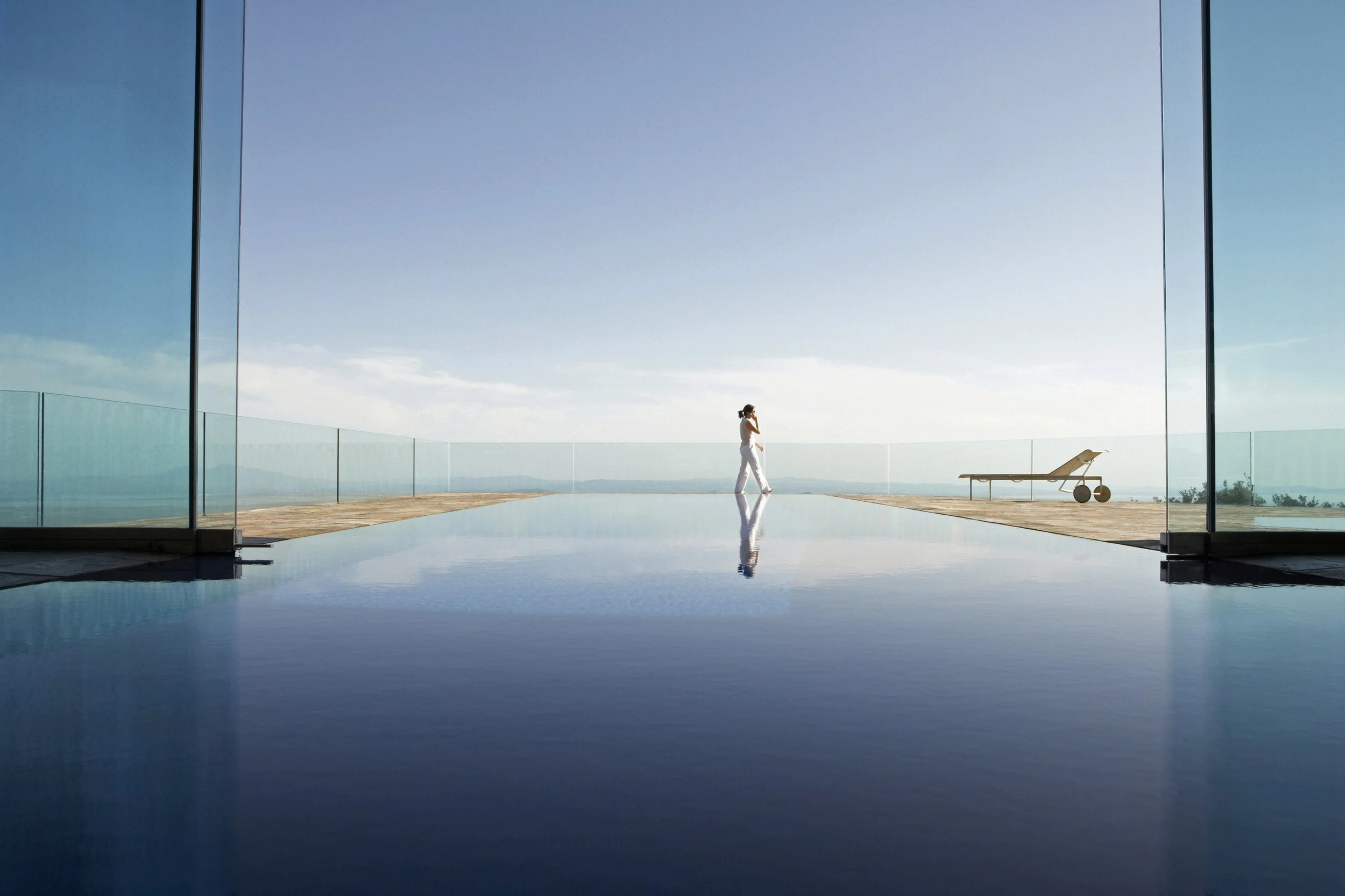 A woman in a white outfit walking along a balcony with glass railings during daytime. There is a lounge chair nearby, and the scene includes a reflection of the woman and the lounge chair in a pool of water. The background shows a clear sky and a distant landscape.