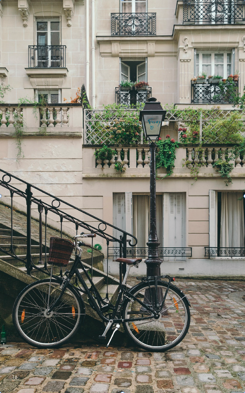 A black bicycle with a wicker basket on the front parked beside a stone staircase in front of a European-style building with balconies and potted plants.