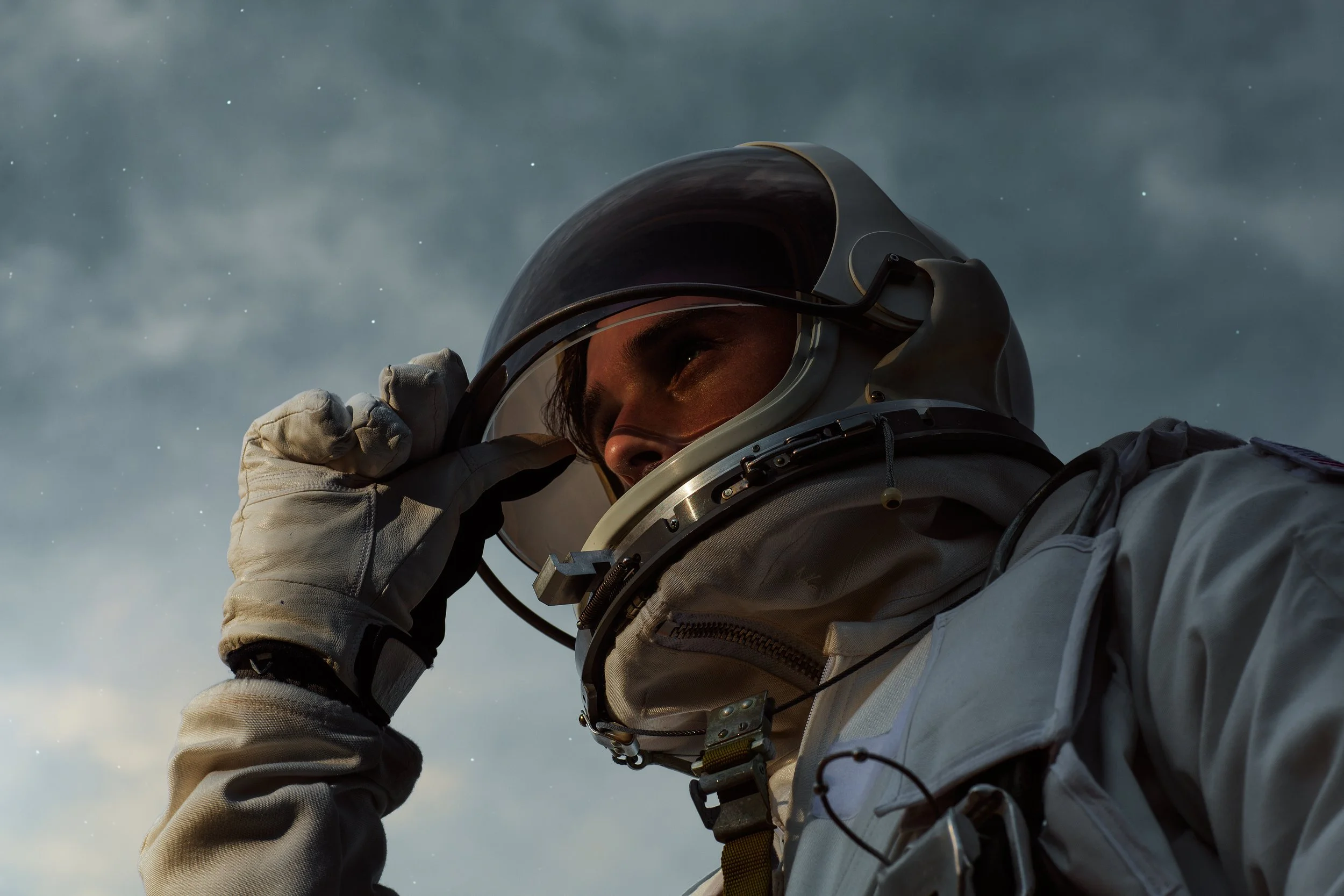 An astronaut adjusting the helmet of their spacesuit outdoors with a cloudy sky in the background.