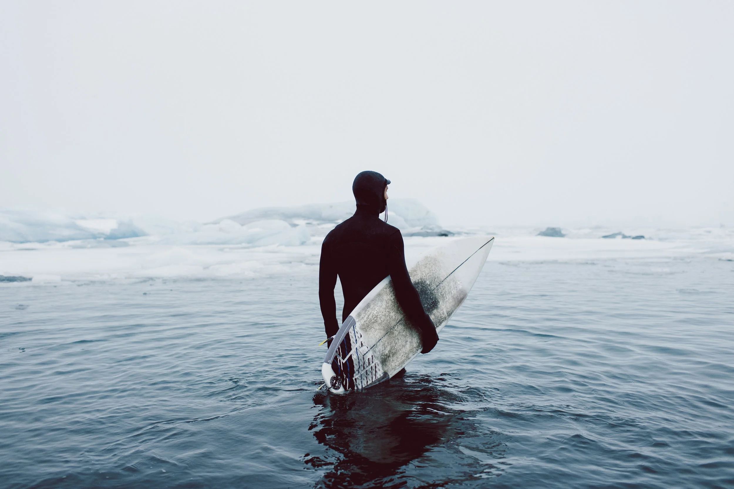 A person wearing a black wetsuit and helmet holding a white surfboard standing in the water near ice-covered terrain, with an overcast sky.