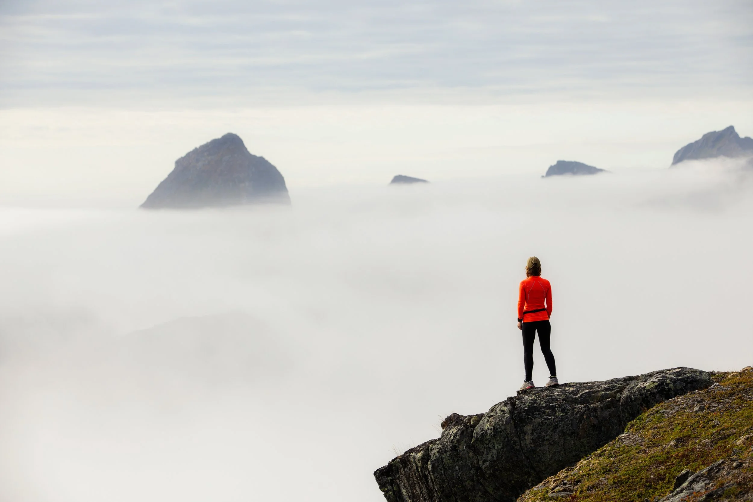 A woman in a red jacket and black pants stands on a rocky outcrop, looking at a mountain range surrounded by clouds and fog with rocky peaks in the distance.