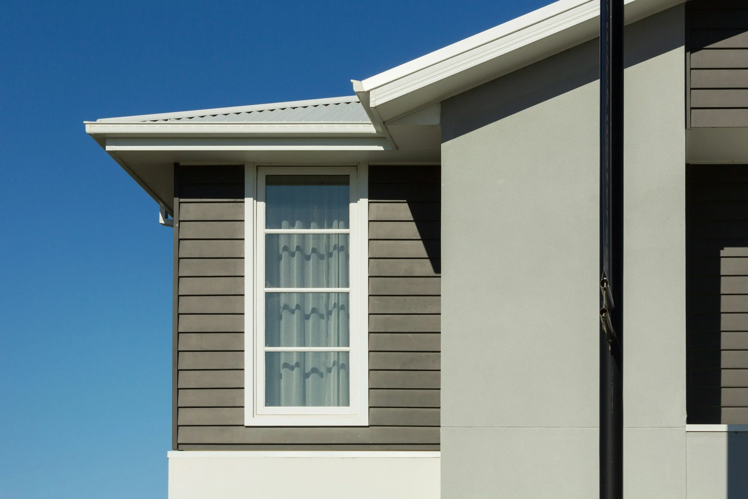 Close-up of a house exterior with tan siding and a white-trimmed window behind a sheer curtain