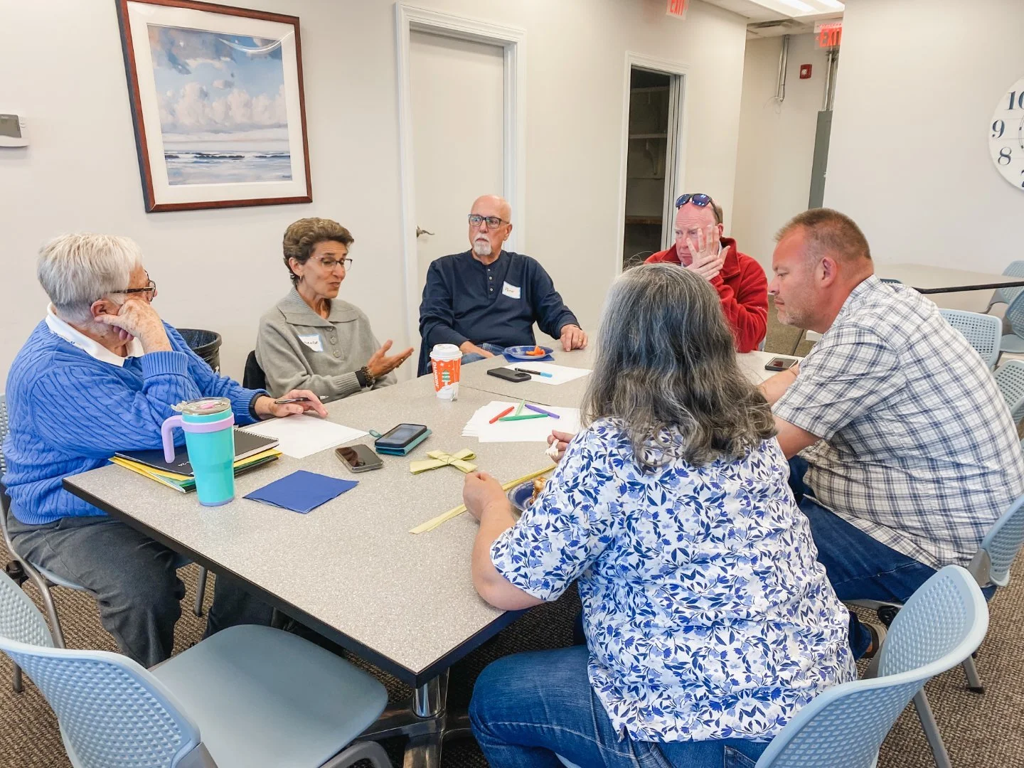 Six people sitting around a conference table engaged in discussion, with notebooks, phones, papers, and snacks on the table in a meeting room.