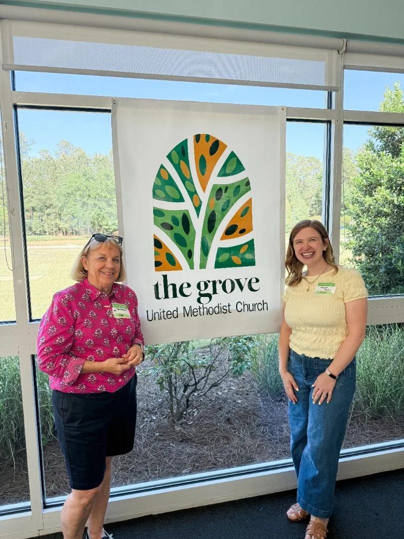 Two women standing inside, smiling, in front of a banner for the Grove United Methodist Church. The banner has a stylized tree logo in green and orange. Outside the window are green trees and shrubs.