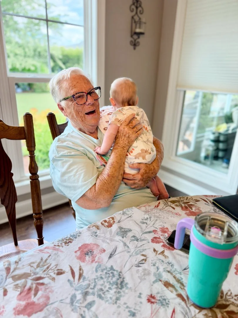 An elderly woman with glasses smiling and laughing while holding a baby at a dining table in a sunlit room.
