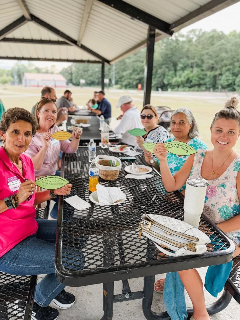Group of people seated at an outdoor picnic table under a pavilion, holding paper leaf-shaped cards, with food and drinks on the table.