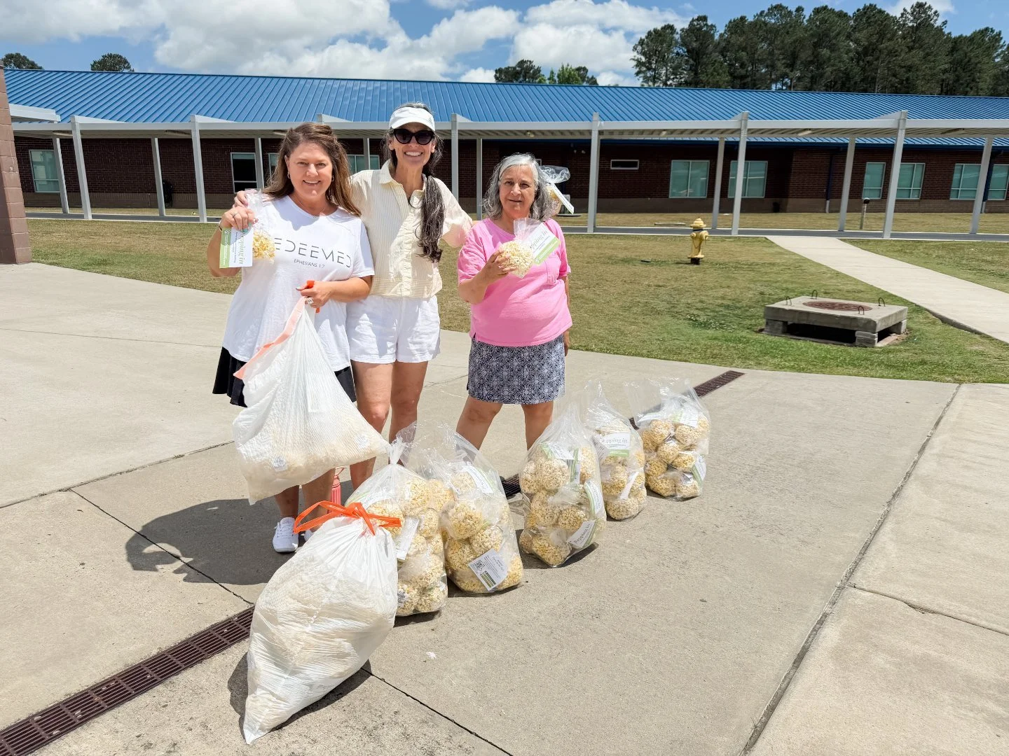 Three women standing outdoors on a sidewalk, holding bags of popcorn, with several large bags of popcorn placed on the ground in front of them. The background features a building with a blue metal roof and a grassy area.