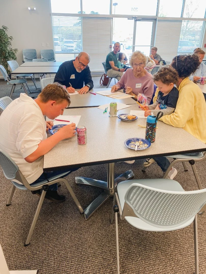 People sitting around a table in a bright room with large windows. They are eating and writing, with papers, snacks, and drinks on the table.