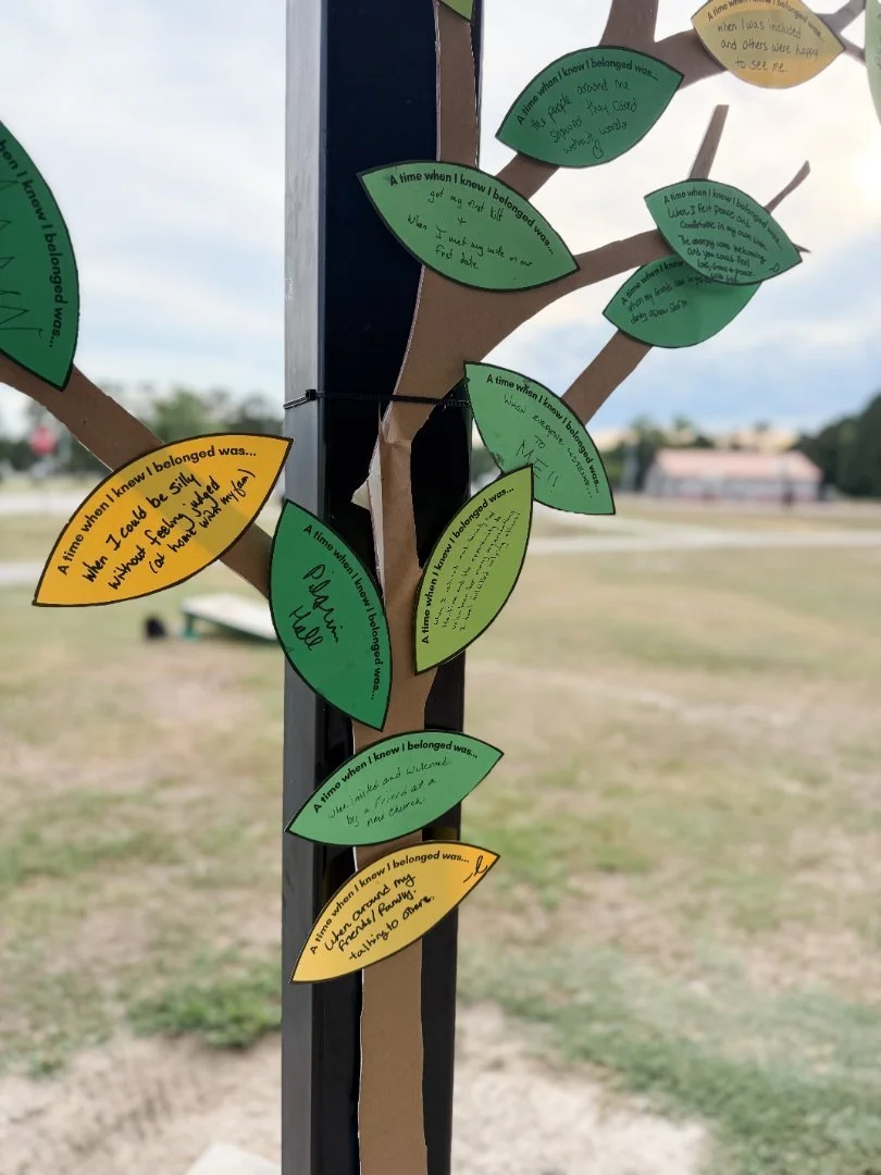 A paper tree display with green and orange paper leaves attached to a cardboard trunk. Each leaf contains handwritten statements about personal feelings of belonging, with some referencing specific moments when the person knew they belonged.