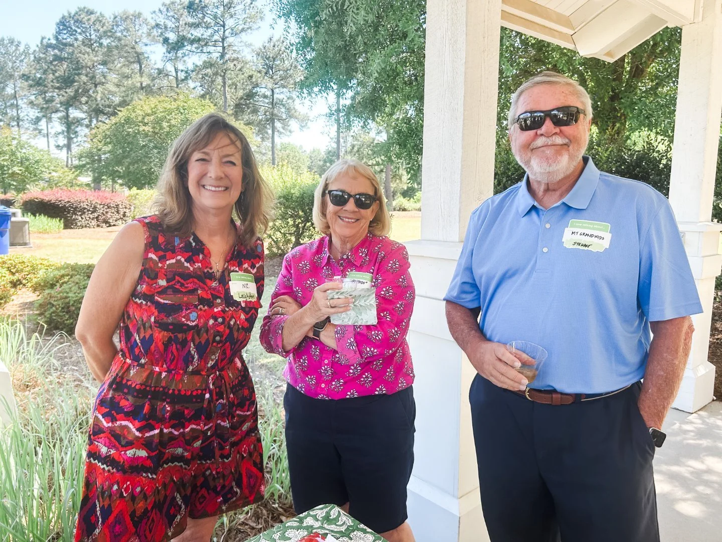 Three smiling adults, two women and one man, standing outdoors on a sunny day at a gathering, wearing sunglasses and name tags, with a garden and trees in the background.