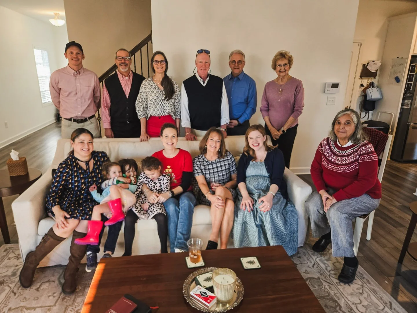 A family gathering in a living room with three women, five men, and four children, smiling for a group photo. The children are seated on a sofa in front, and the adults stand behind them. The room has a cozy, lived-in feel with a wooden coffee table and a white wall.