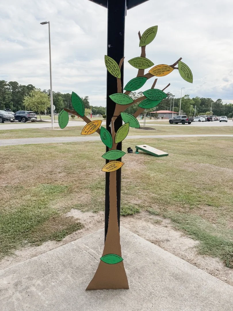 A decorative tree with paper leaves, placed outdoors on a sidewalk near a parking lot and grassy area.