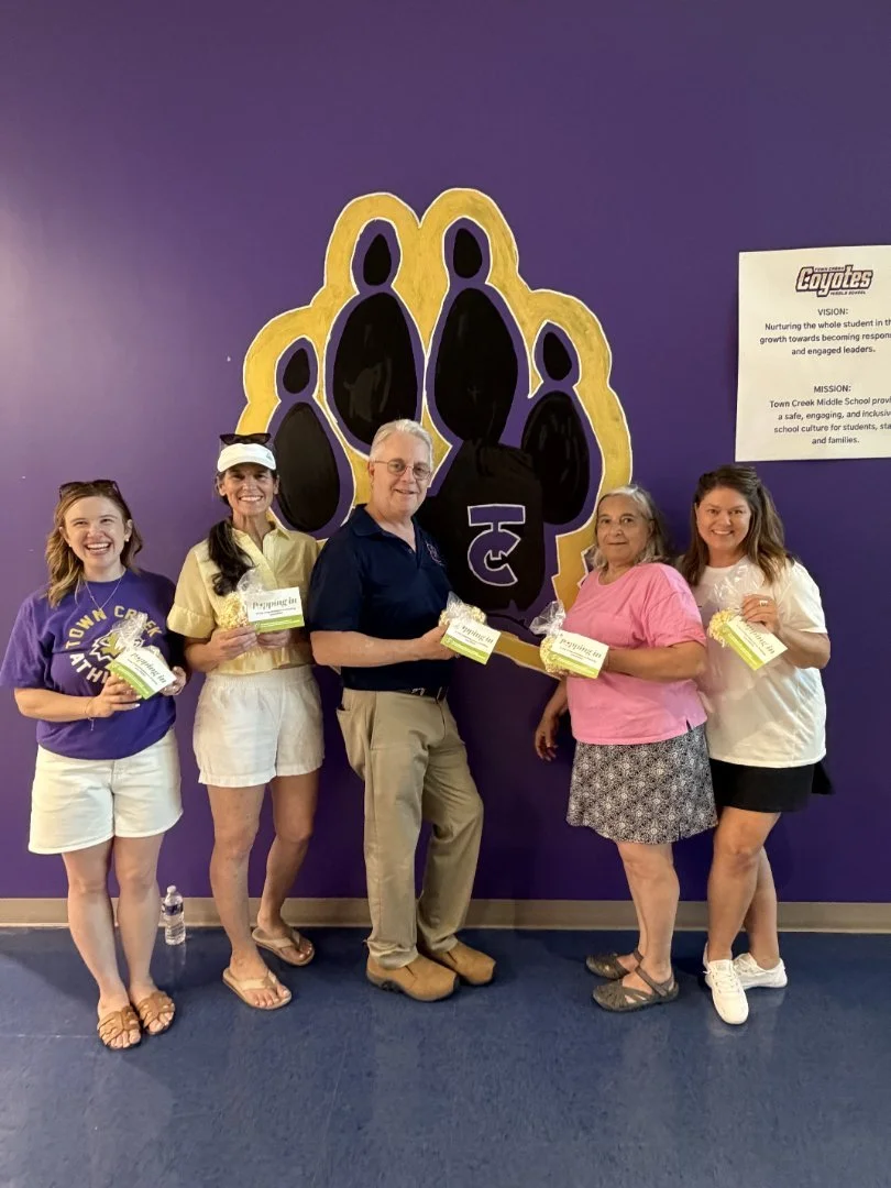 Five women standing in front of a purple wall with a large bear paw print logo, holding packs of popcorn, smiling at the camera.