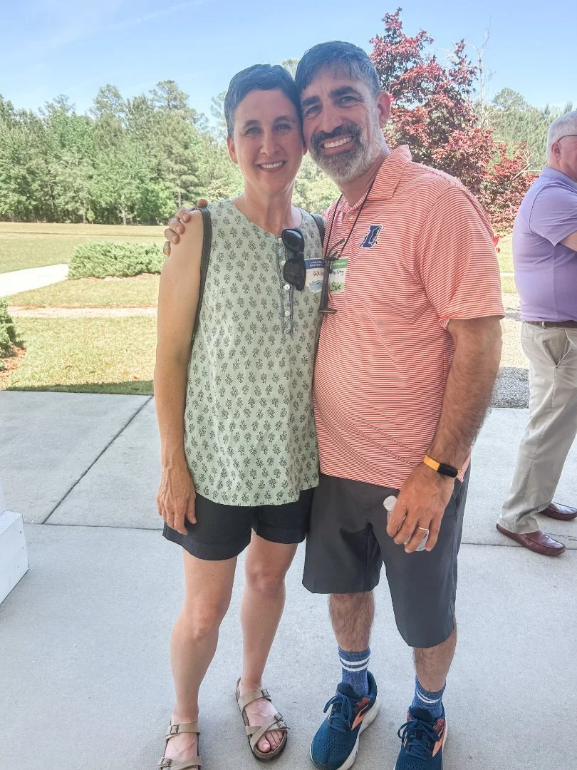 A man and woman smiling outdoors on a sunny day, standing close together with arms around each other's shoulders. The woman is wearing a sleeveless patterned top, black shorts, and sandals. The man is wearing a striped polo shirt, shorts, sneakers, and colorful socks. There is greenery and a tree with red leaves in the background.