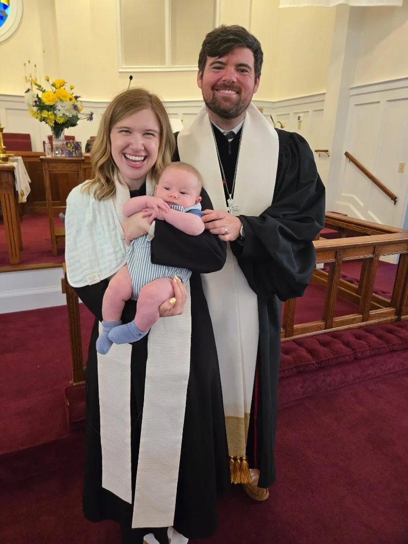 A woman, a man in a church robe, and a baby during a baptism ceremony in a church with wooden pews and floral arrangements.