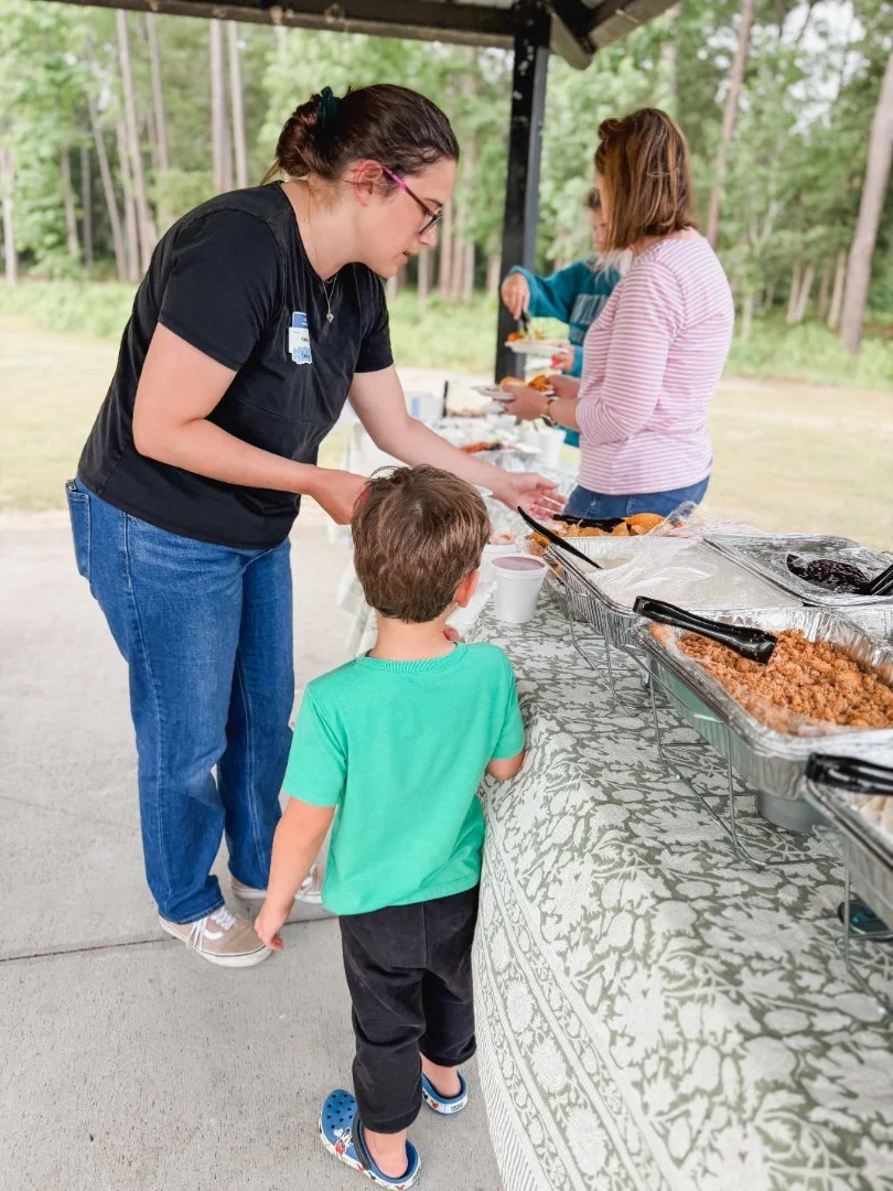 A woman serving food at an outdoor buffet table while a young boy in a green shirt and black pants watches. In the background, two women are also serving food, with trees and grass visible.